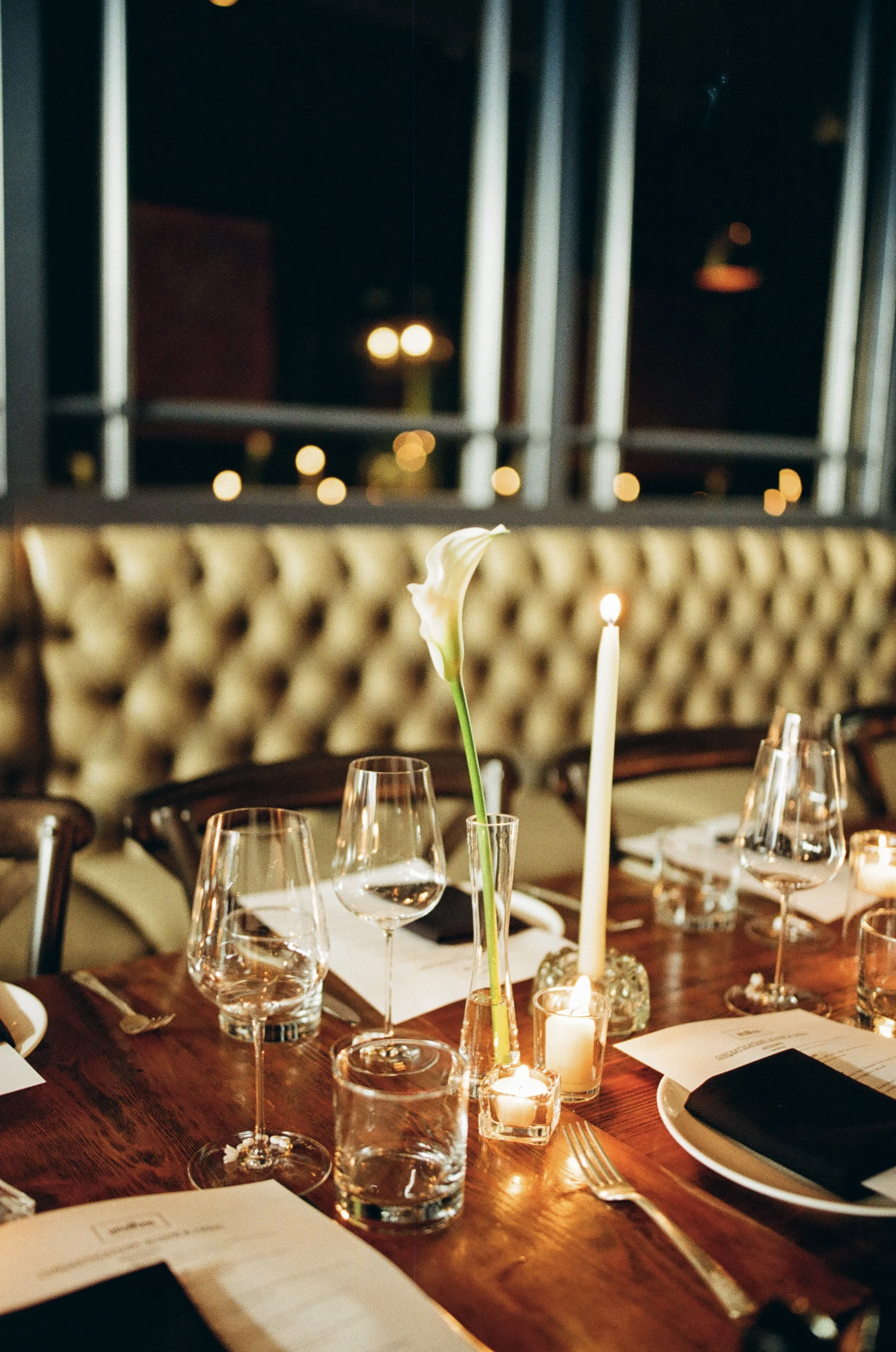 Table set for dinner with wine glasses, candles, a white flower, paper menus, plates, and silverware, in a dimly lit restaurant with a tufted upholstered wall and large windows in the background.