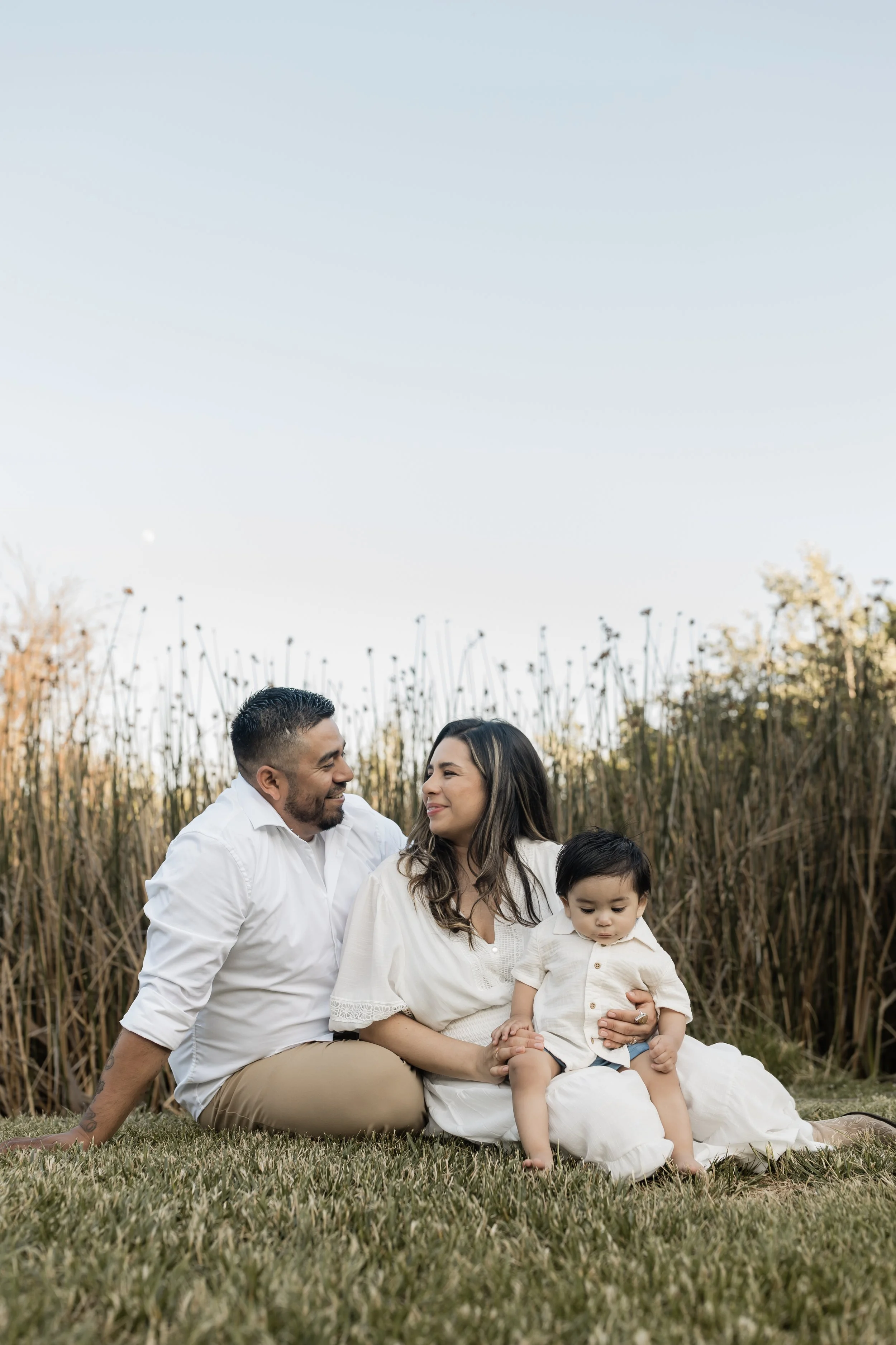 Family of four sitting outdoors on grass during daytime, with tall plants and clear sky in the background. The father and mother are smiling at each other, and their young son is sitting on the mother’s lap, looking down.