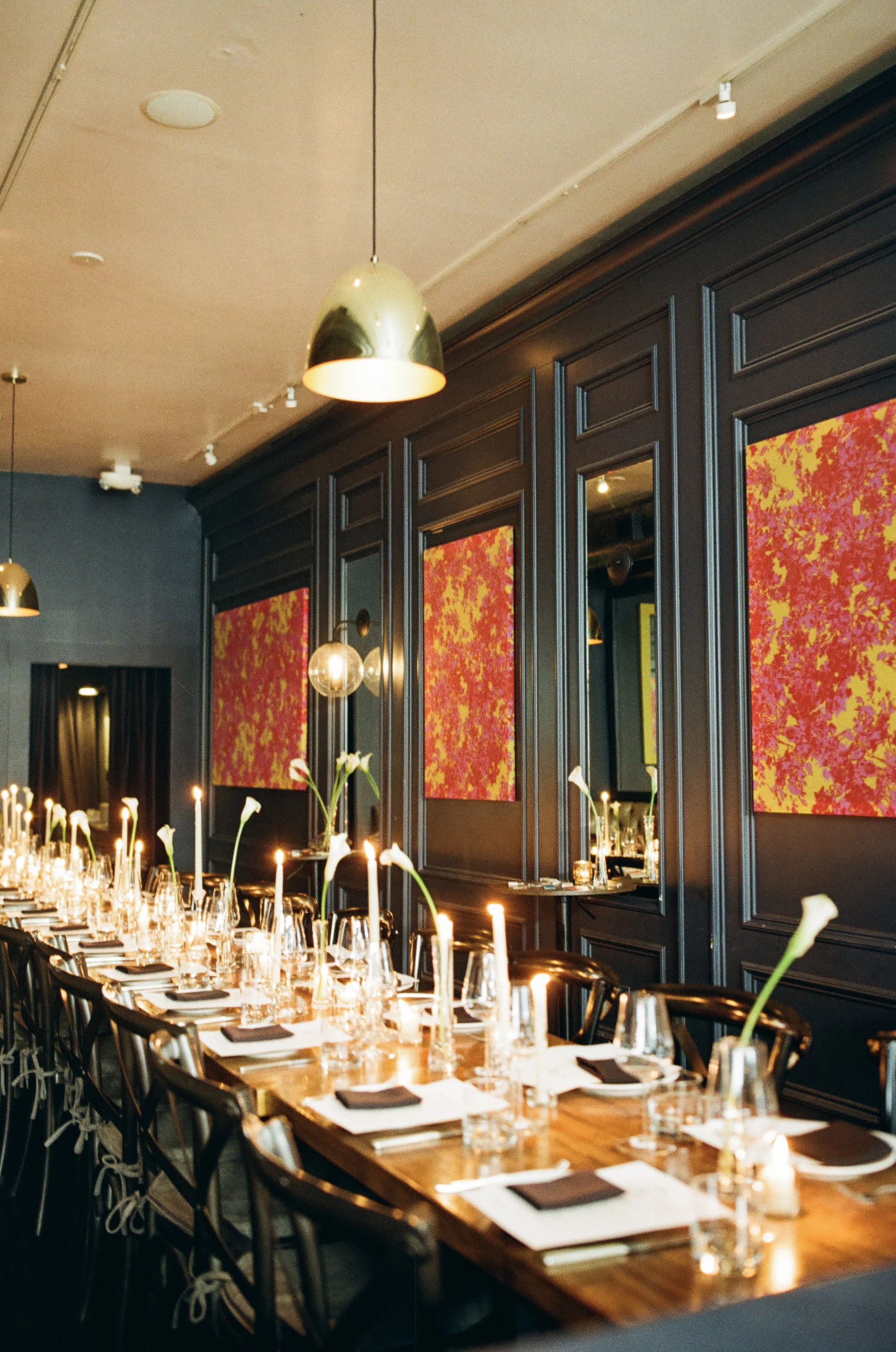 A formal dining setup at a restaurant with a long wooden table, surrounded by black chairs, set with wine glasses, candles, and napkins. The room features dark paneled walls decorated with abstract red and orange artwork and warm pendant lighting.