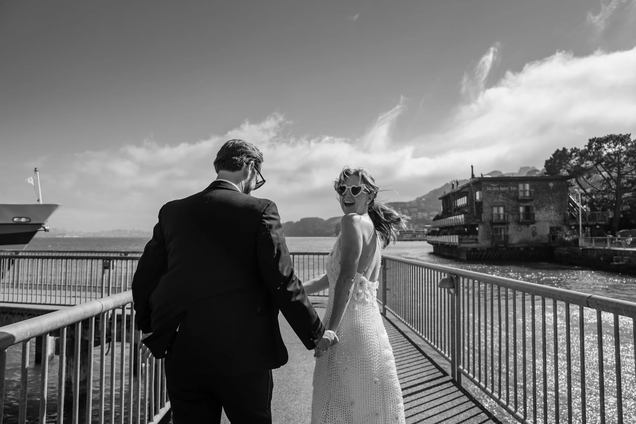 A black-and-white photo of a man and woman holding hands by a waterfront, with the woman wearing heart-shaped sunglasses and both dressed nicely, possibly for a special occasion.