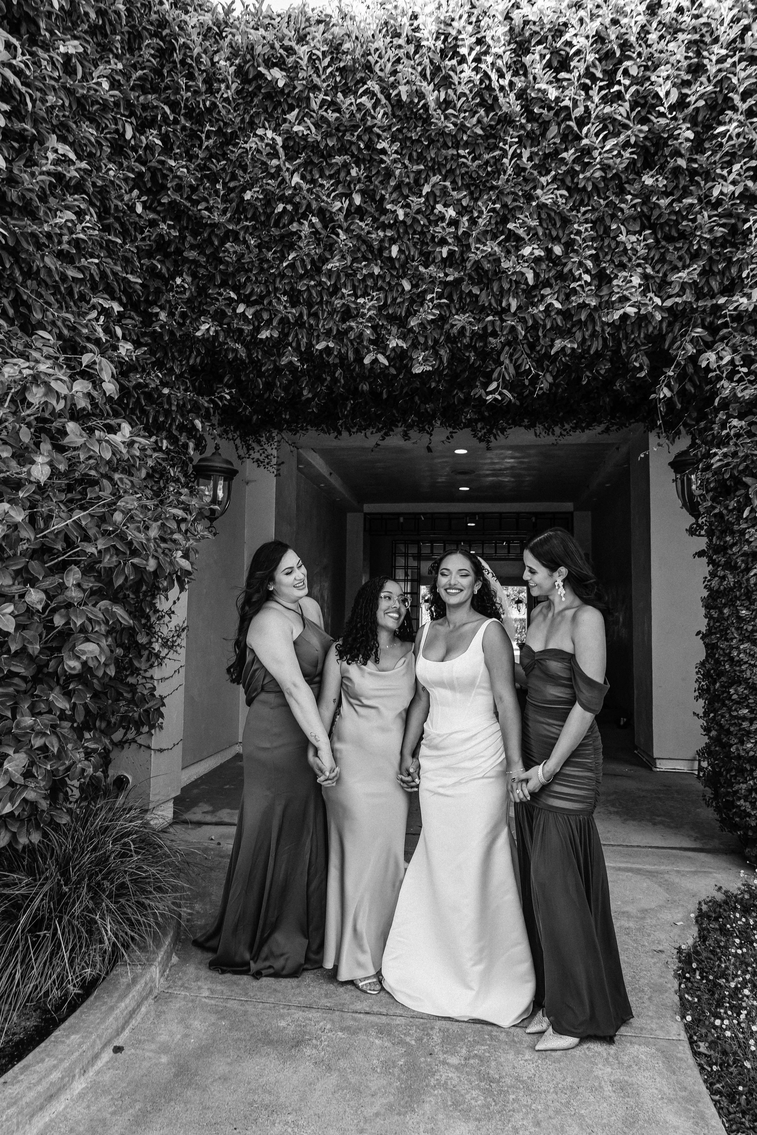 Black and white photo of five women dressed in formal gowns, standing close together outdoors under a large leafy arch, smiling and holding hands.
