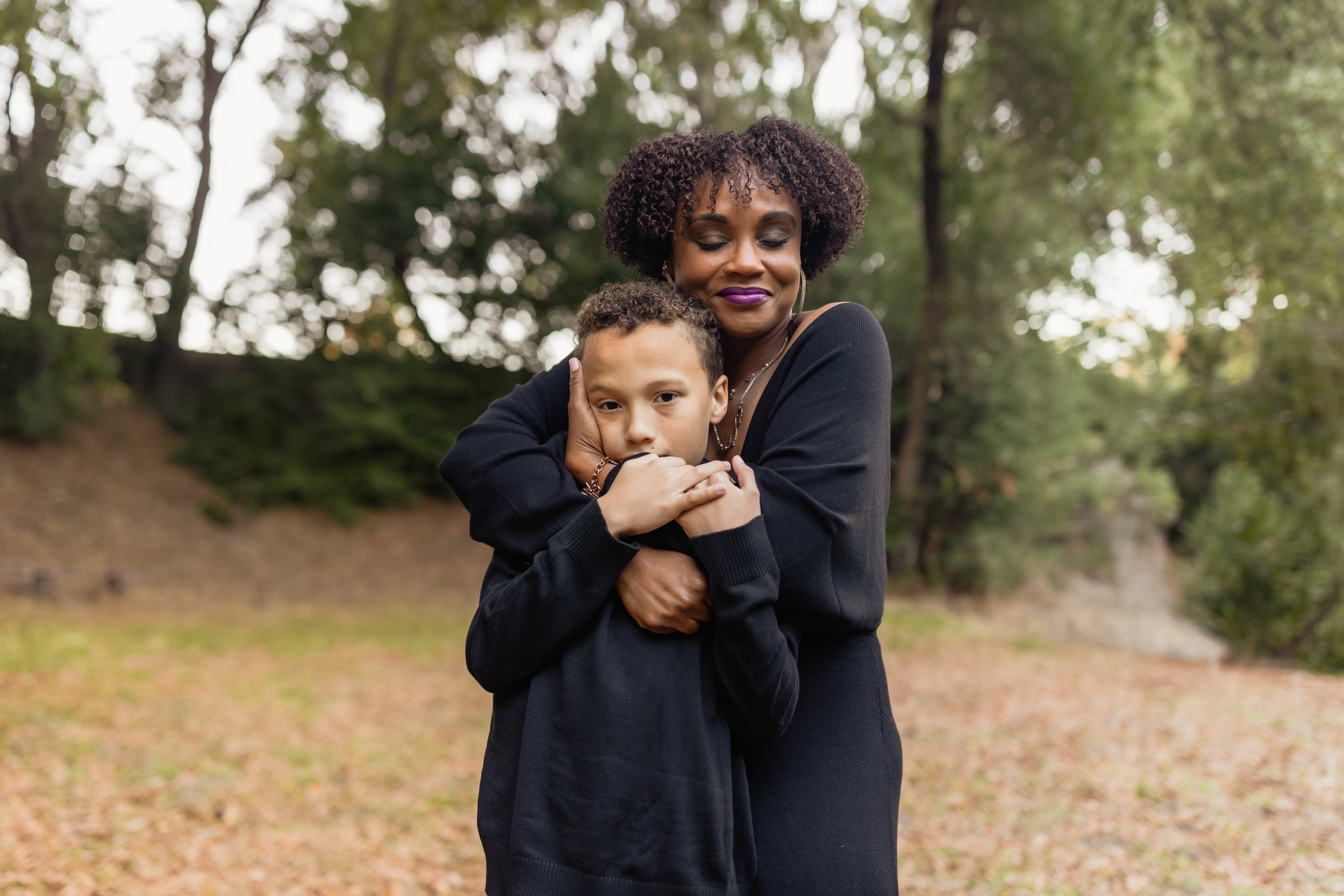 An African American woman with curly hair and purple lipstick hugging a young boy with short curly hair outdoors in a park with trees in the background.