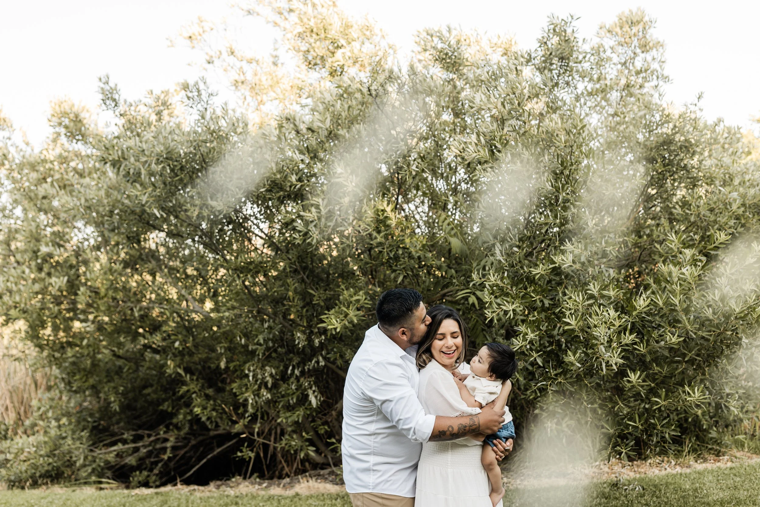 A family of three, including a man, woman, and young girl, standing outdoors in front of large green trees. The man is holding the girl and they are smiling.