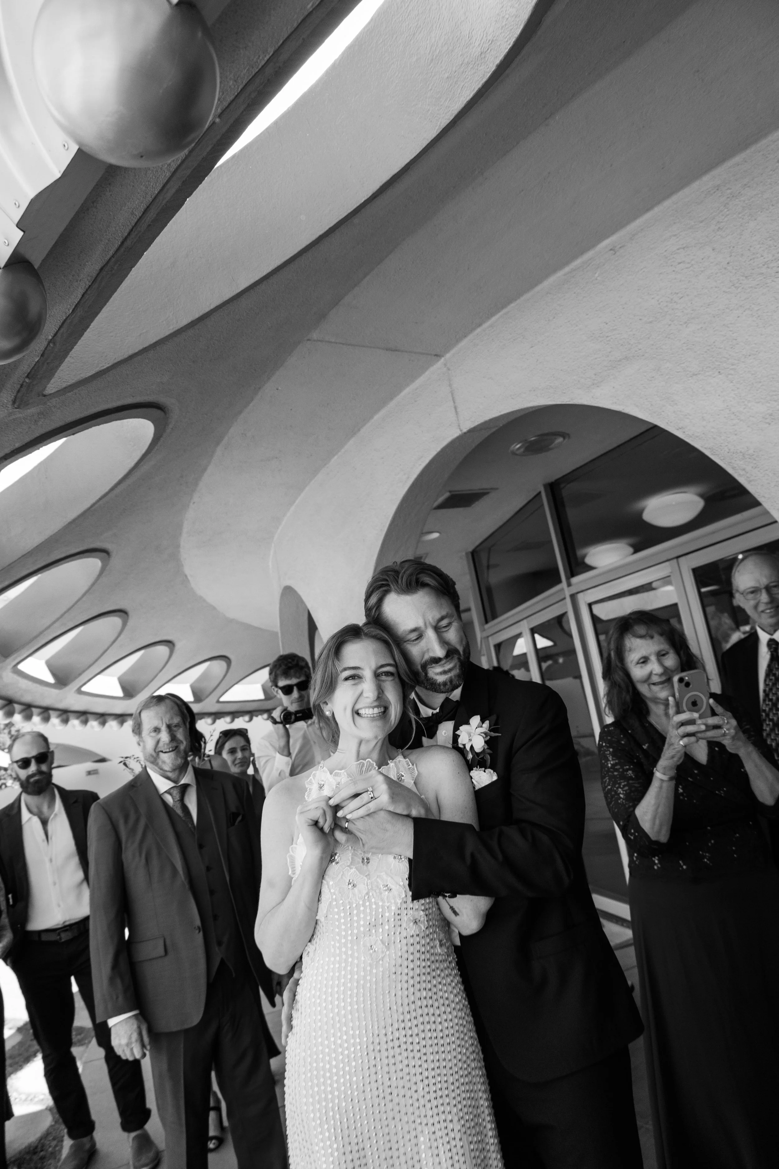 A group of people at a wedding, with a bride and groom at the center. The crowd is smiling and taking photos, with a modern, curved building with circular windows in the background.