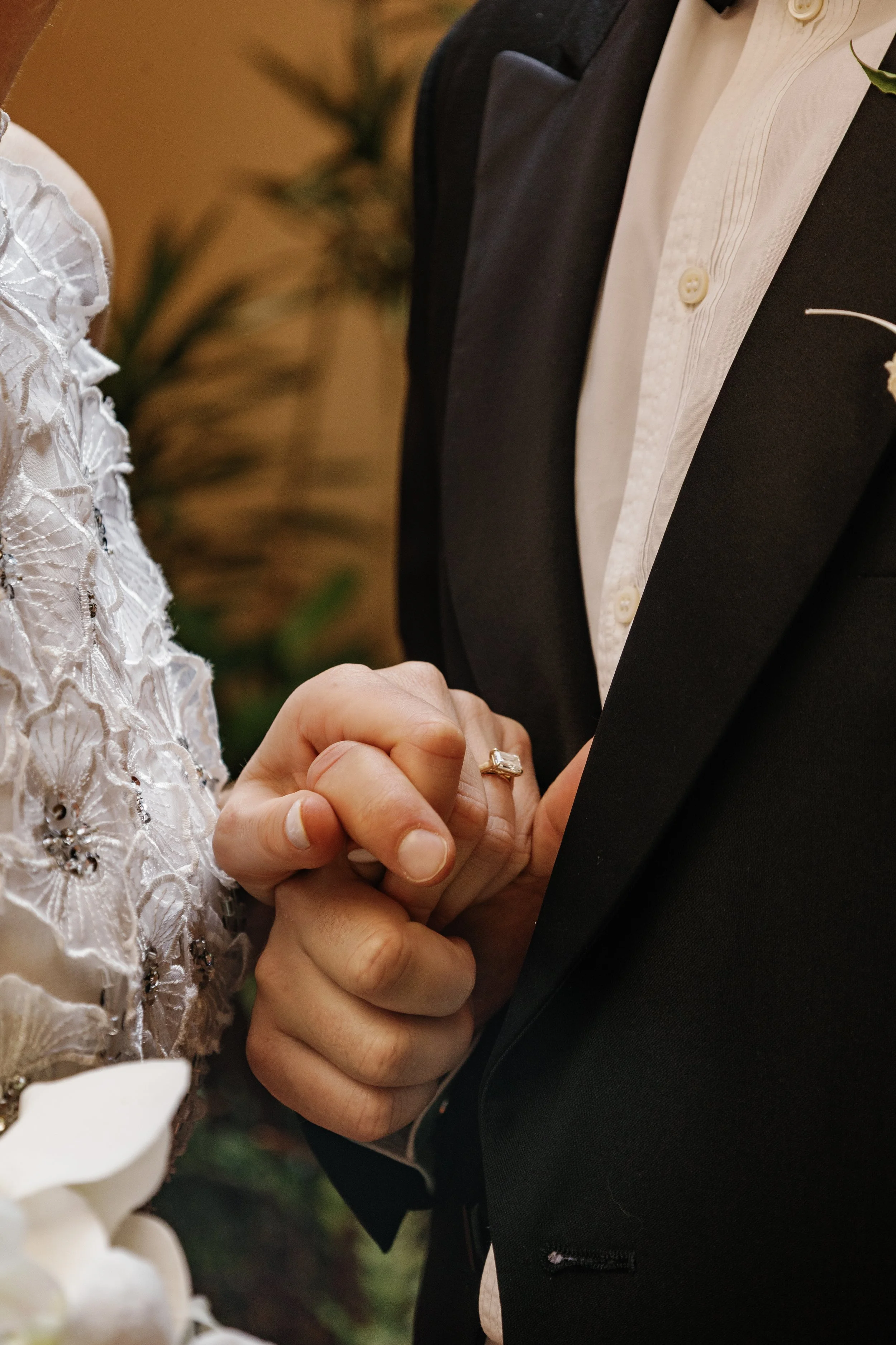 A close-up of a couple holding hands during their wedding ceremony. The bride is wearing a white lace dress, and the groom is dressed in a black tuxedo with a white shirt and a boutonnière. The focus is on their hands with the bride's engagement ring