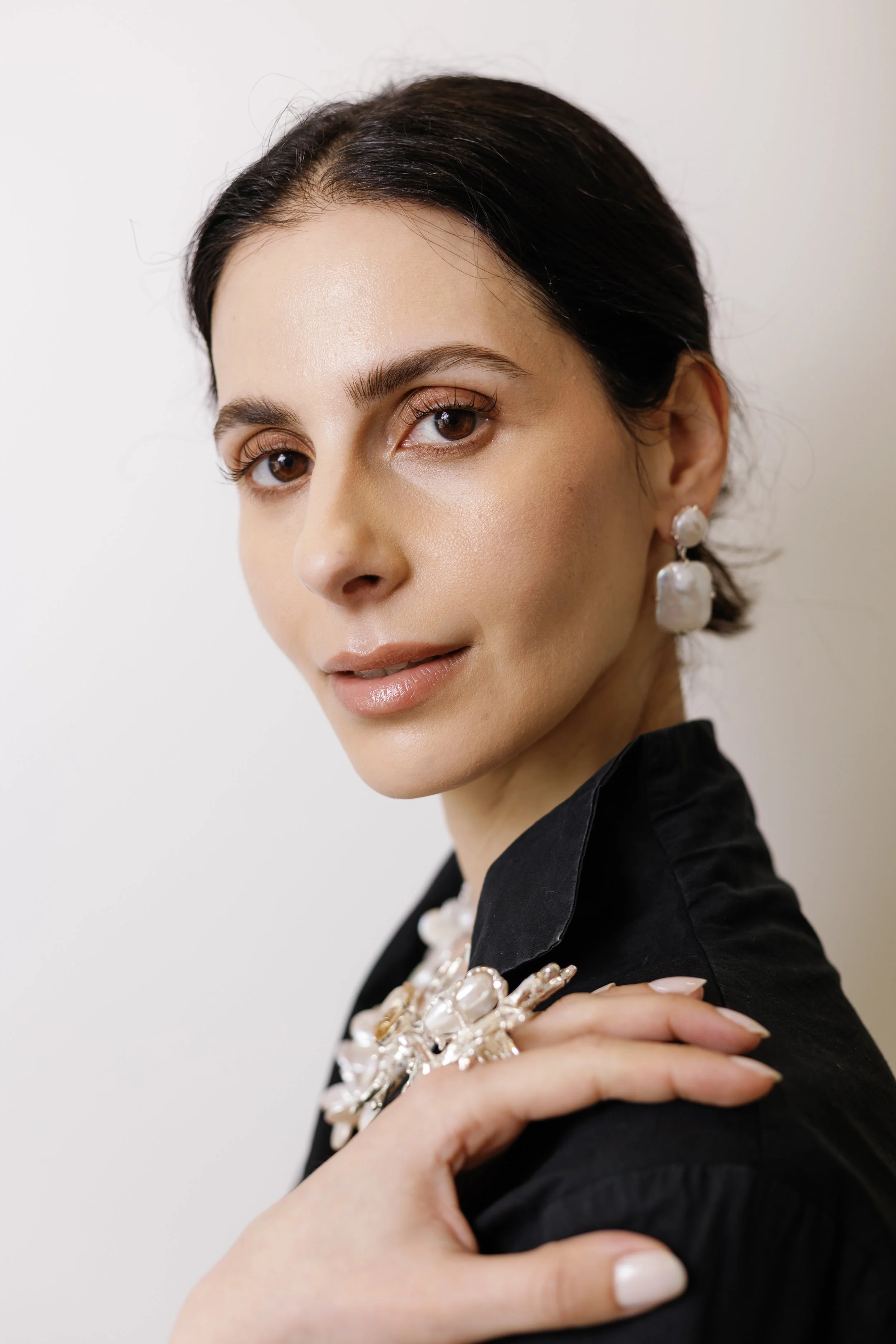 Close-up of a woman with dark hair, wearing pearl earrings and matching jewelry, dressed in a black shirt, standing against a white background.