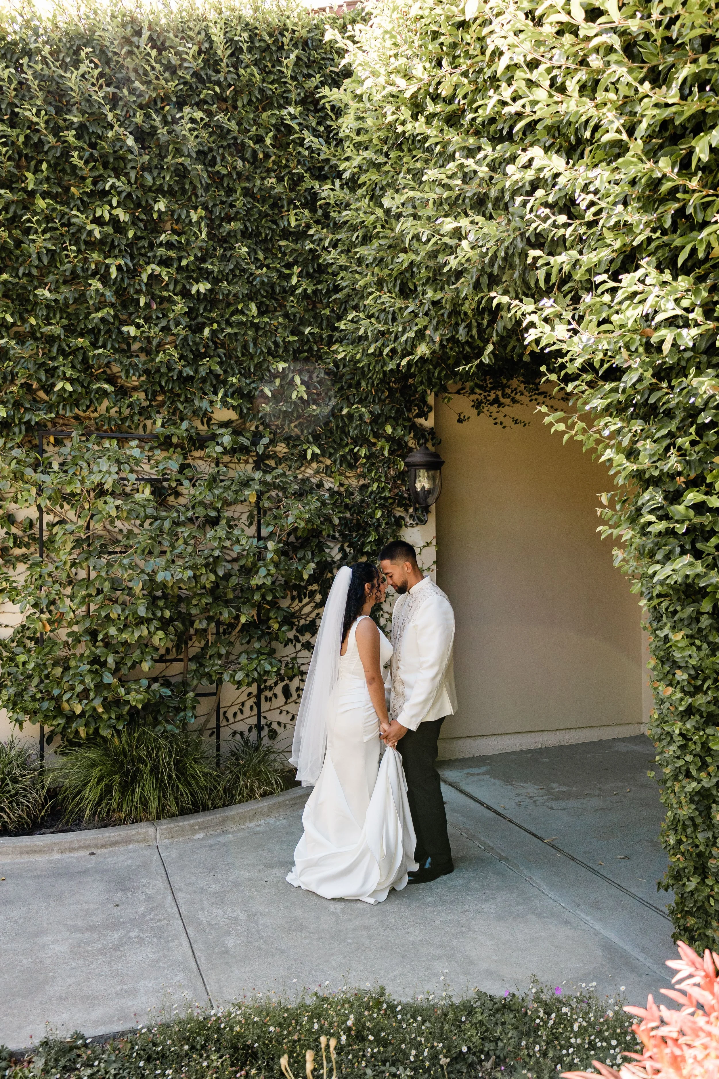 A bride and groom are standing close together outdoors, holding hands, in a garden area surrounded by lush green bushes, during their wedding ceremony.