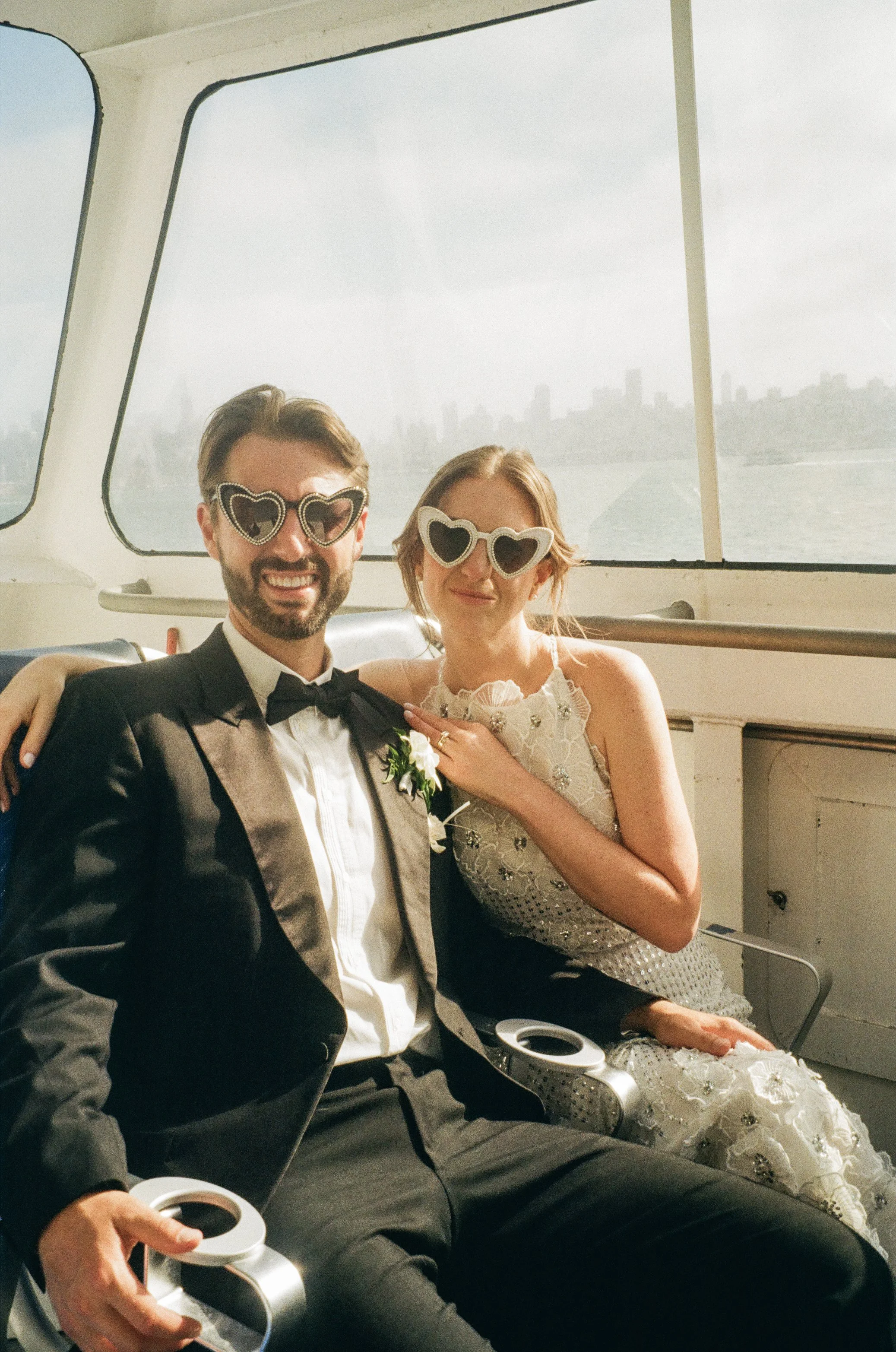 A smiling man and woman in formal attire, wearing heart-shaped sunglasses, sitting on a boat with city skyline visible through the window behind them.