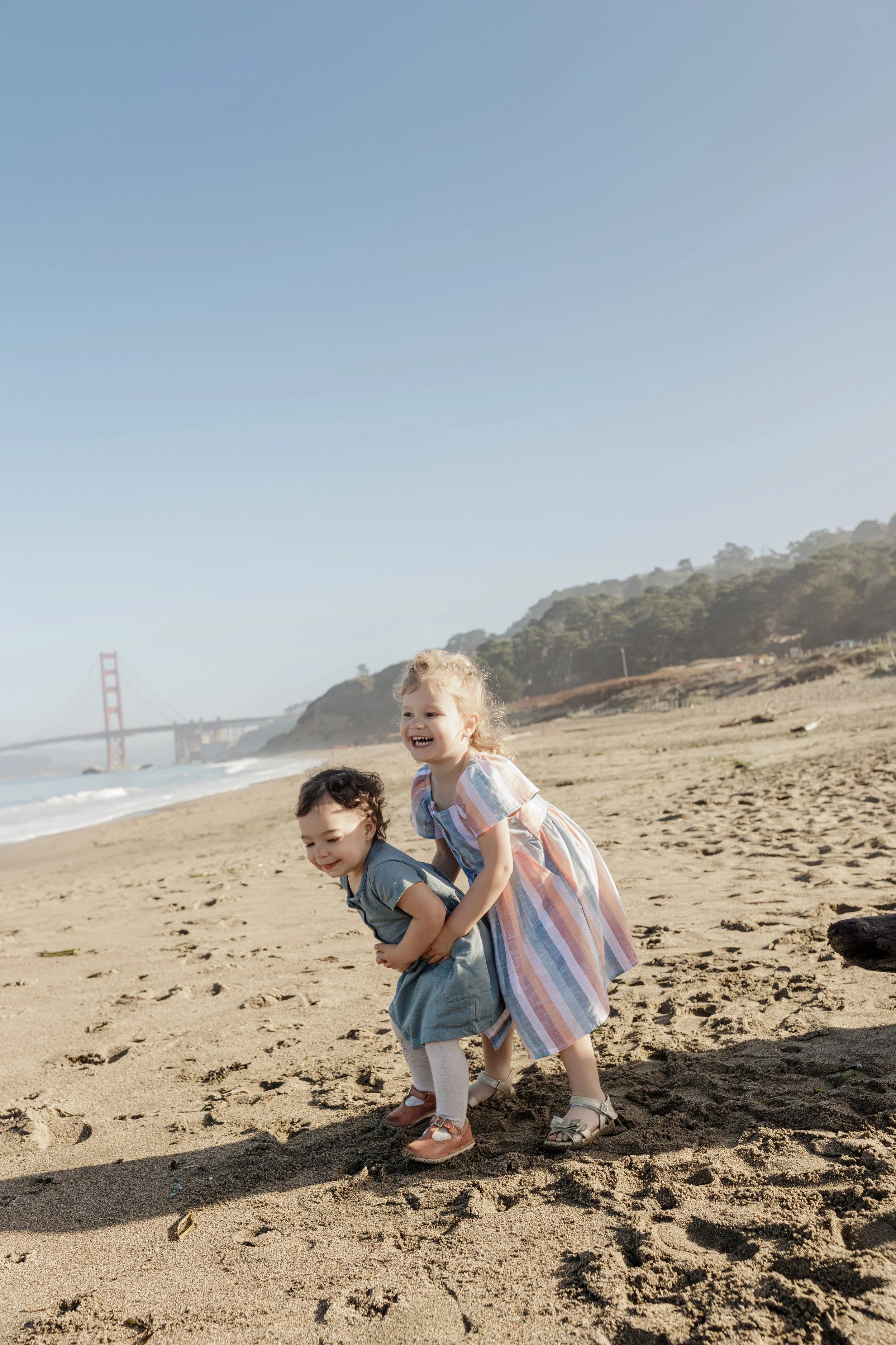Three young children playing and smiling on a sandy beach near water, with the Golden Gate Bridge and green hills in the background.