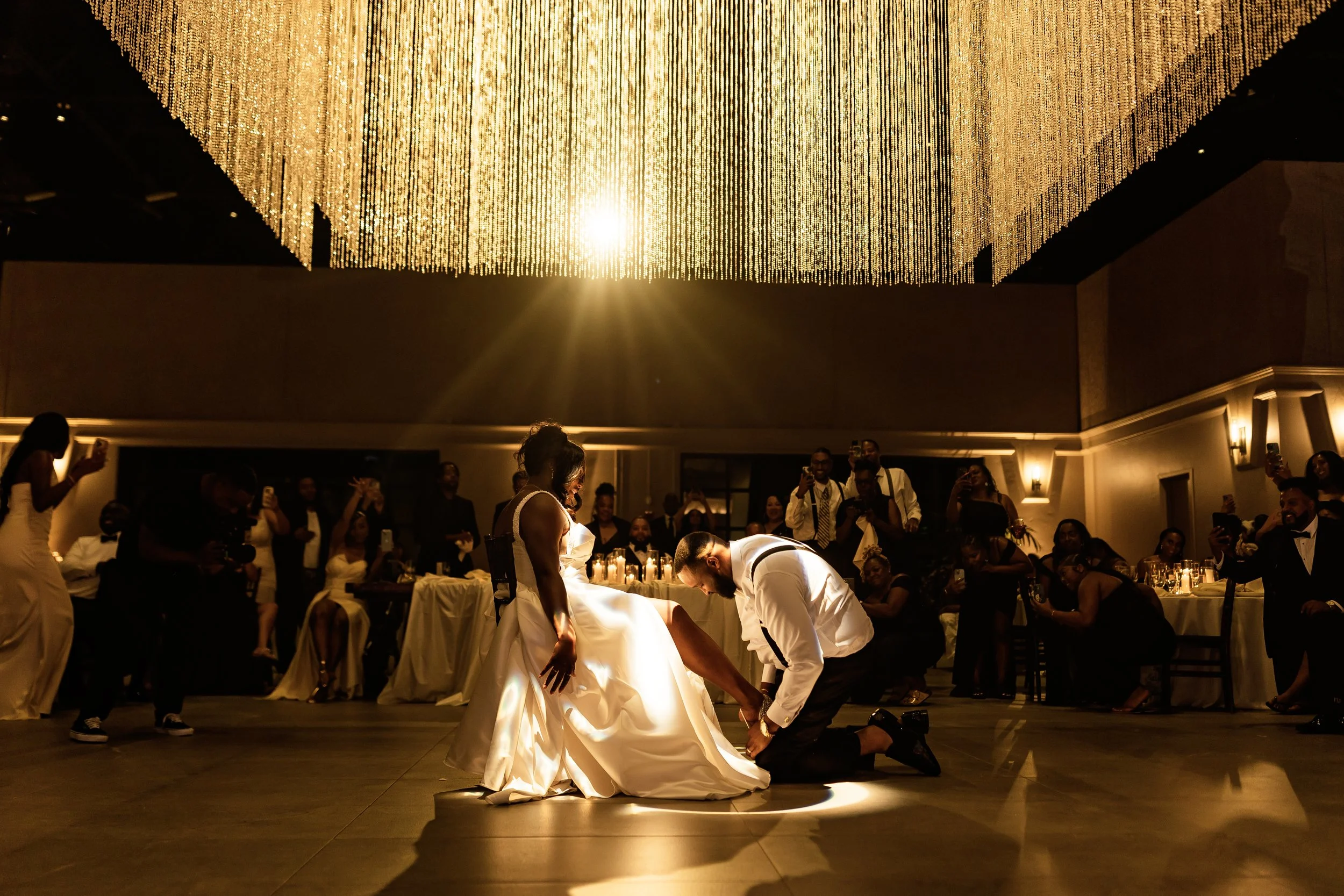 A wedding reception with a chandelier hanging above, guests taking photos, and the bride and groom dancing with guests gathered around.
