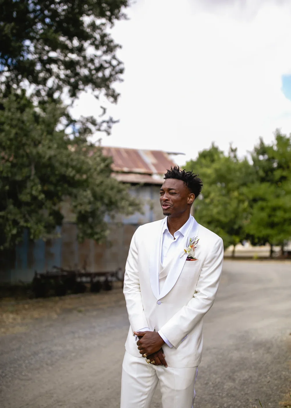 A man in a white suit with a boutonniere on his left lapel standing outdoors on a dirt road, with trees and an old building with a rusted roof in the background.