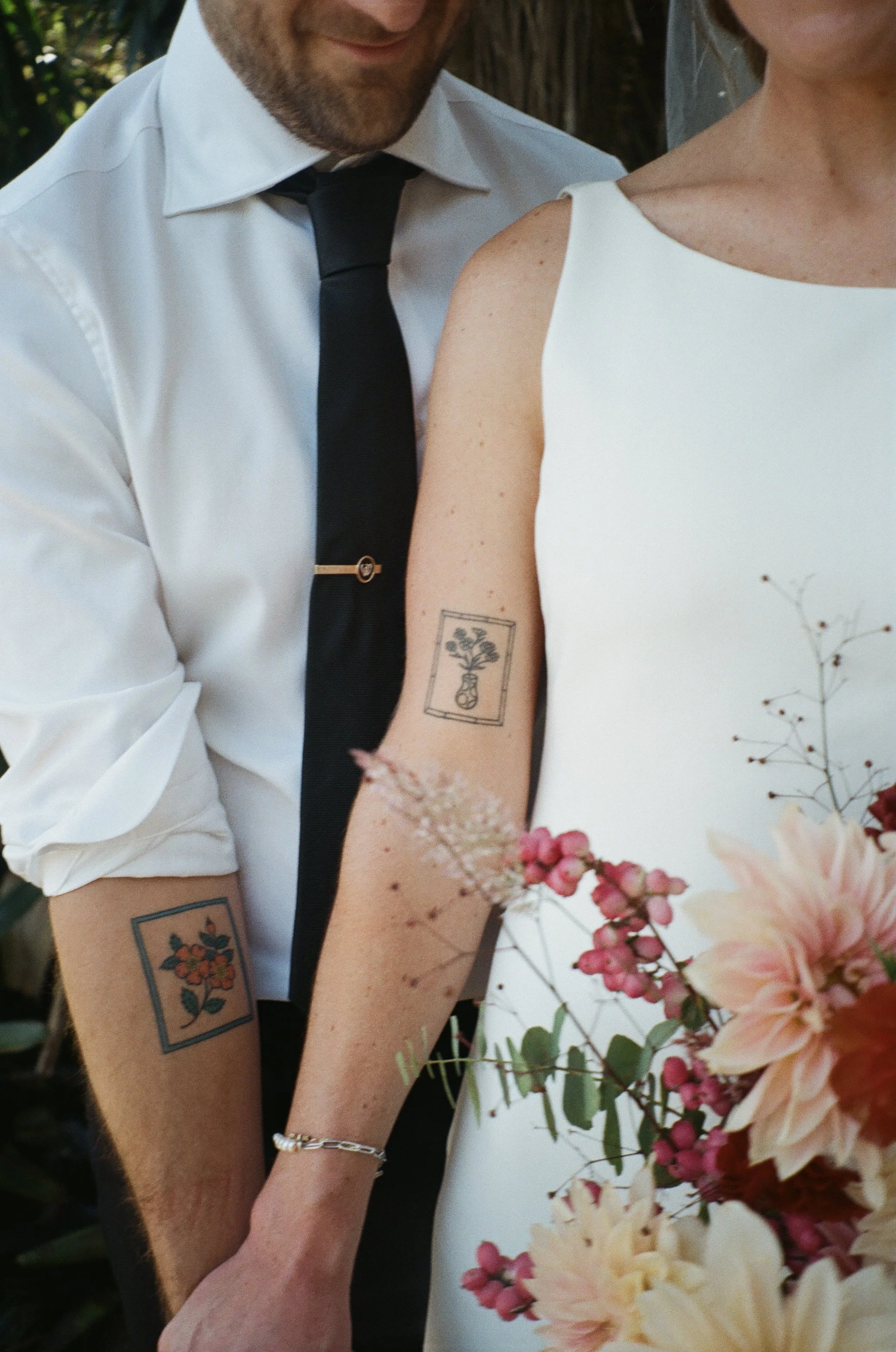 A couple is posing closely together, with the man wearing a white shirt, black tie, and visible tattoos on his arm, while the woman is wearing a white dress with floral designs. They are holding hands amidst a background of lush greenery and colorful