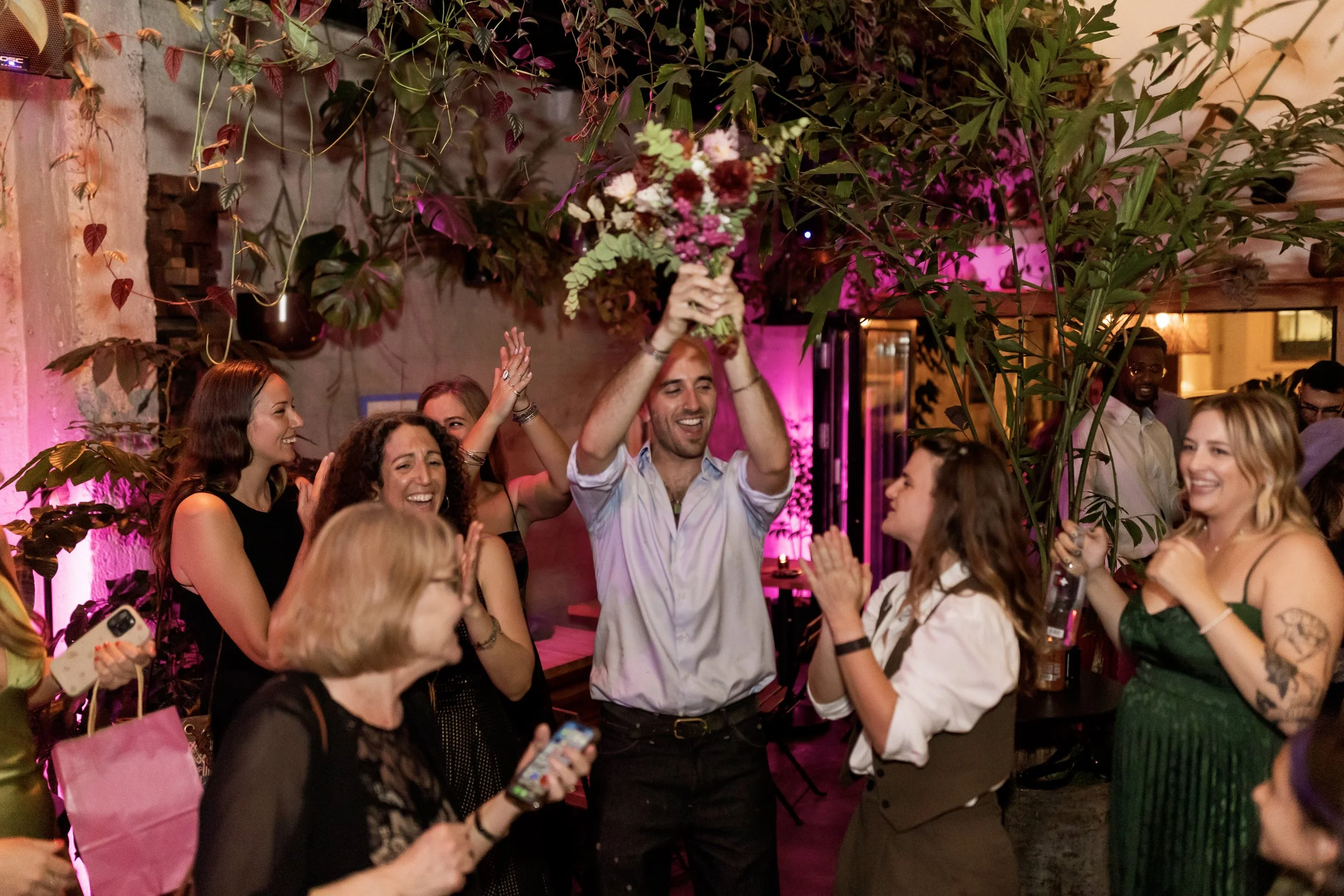 People celebrating at a party, with a man holding a bouquet of flowers above his head, surrounded by smiling women and pink lighting, in a decorated indoor venue with plants.