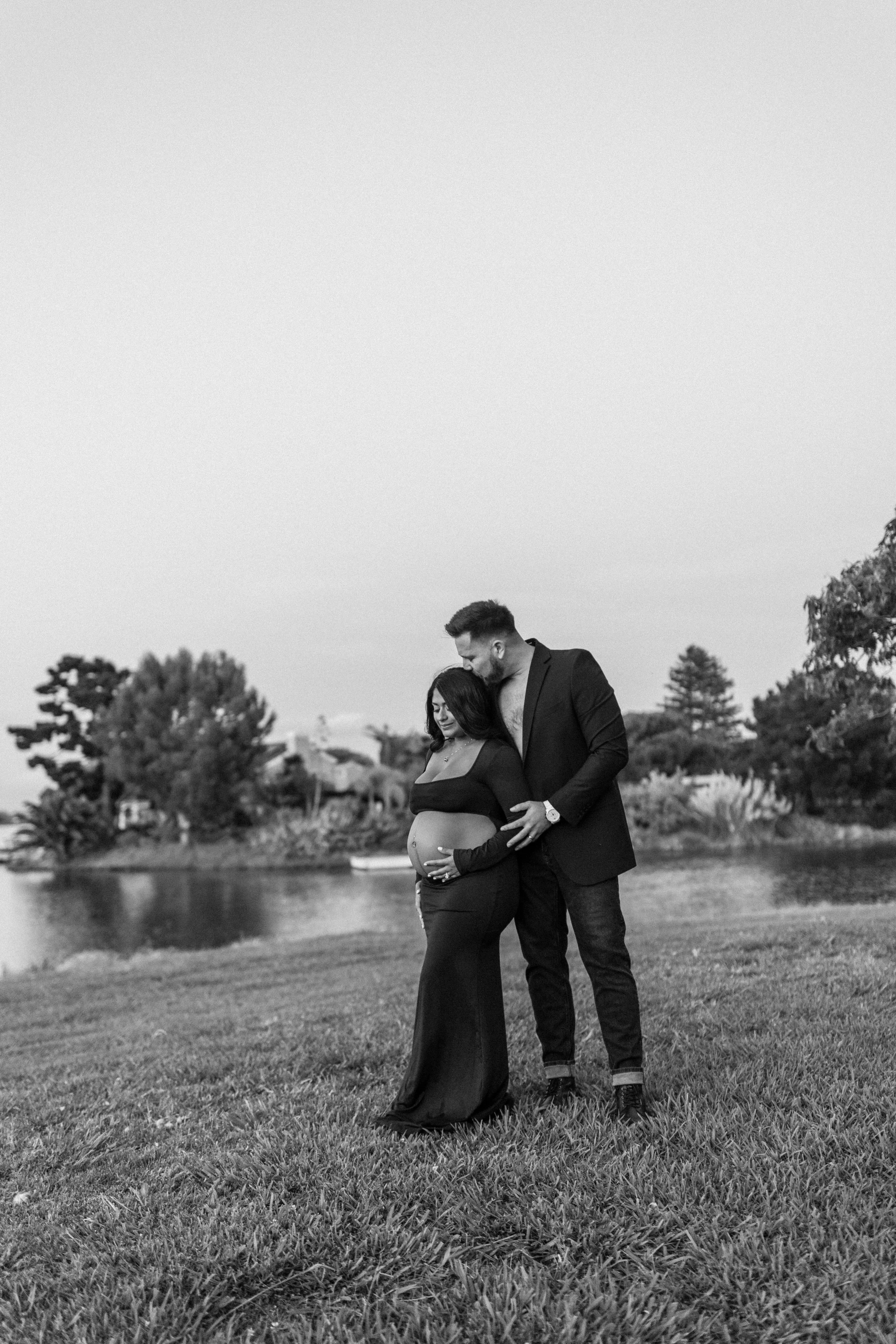 A black and white photo of a pregnant woman and a man standing by a river in a park, with trees in the background. The man is gently kissing the woman's forehead, and they both have their eyes closed. The woman is wearing a long dress that shows her 