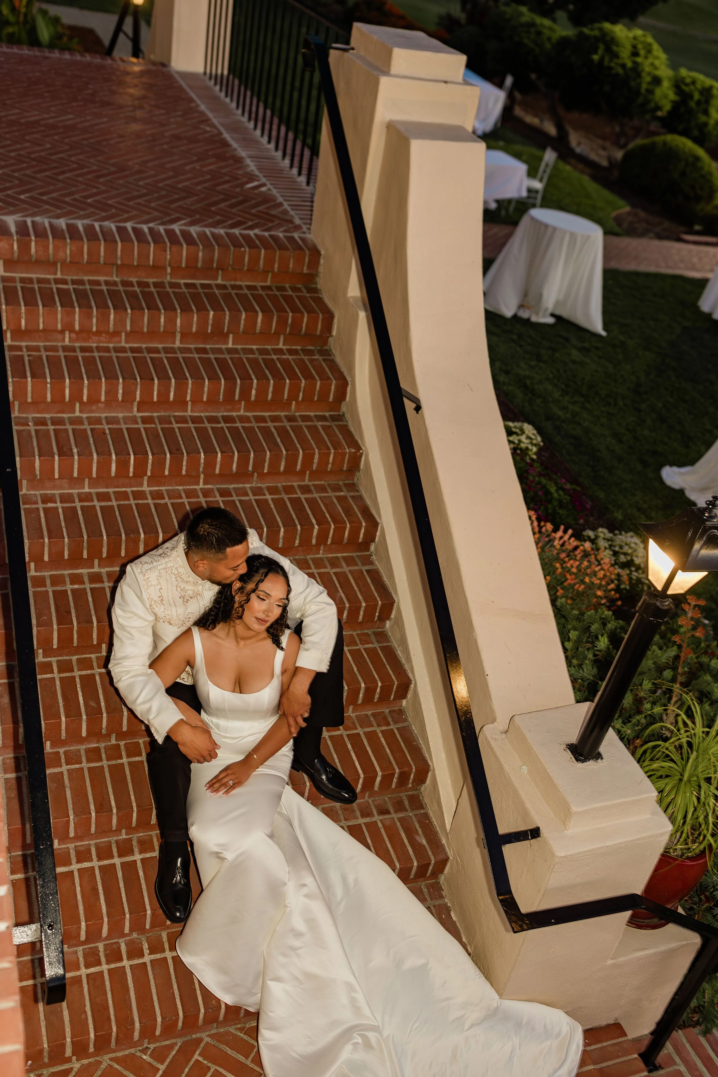 A bride and groom lying on brick stairs, with the groom embracing the bride from behind. The bride has dark curly hair and is wearing a white wedding dress with a low neckline. The groom has short dark hair and is wearing a white jacket with gold emb