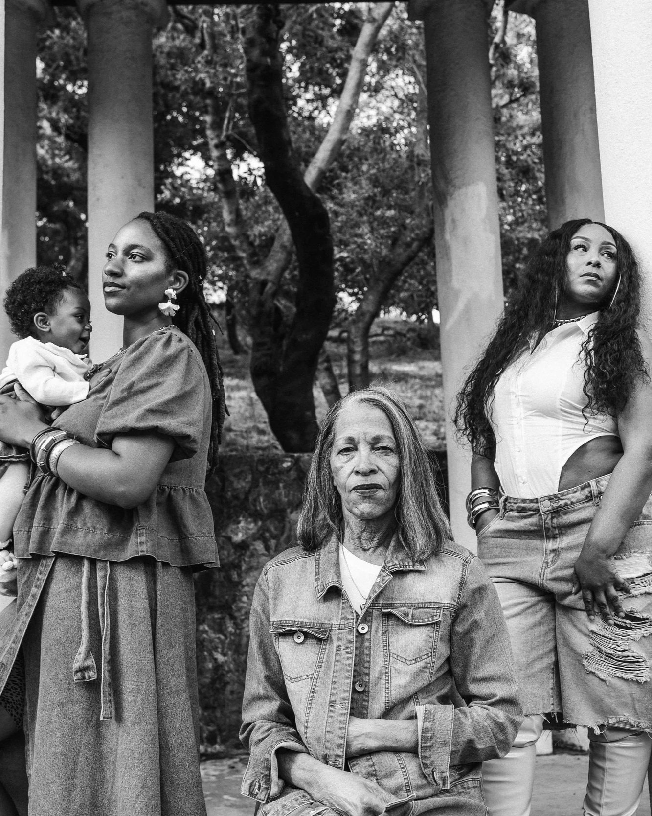Black and white photo of four women and a child outdoors among trees. One woman holds a child, another woman has long, curly hair, and a third woman has shoulder-length gray hair. The woman with gray hair is seated, while the others stand around her.