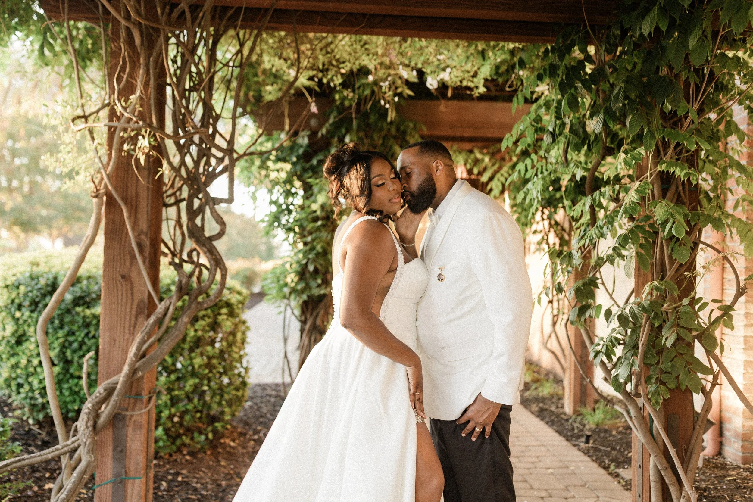 A couple dressed in wedding attire sharing an intimate moment beneath a vine-covered wooden archway in a garden setting.