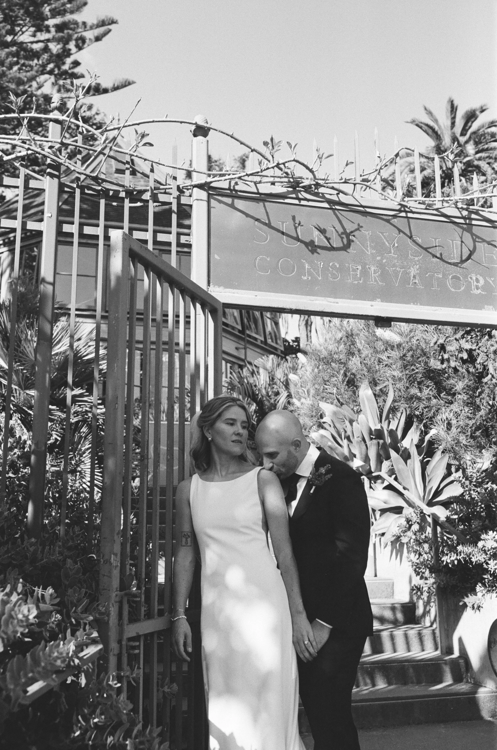 A black and white photo of a bride and groom during their wedding ceremony, standing under an archway with trees and a building in the background.