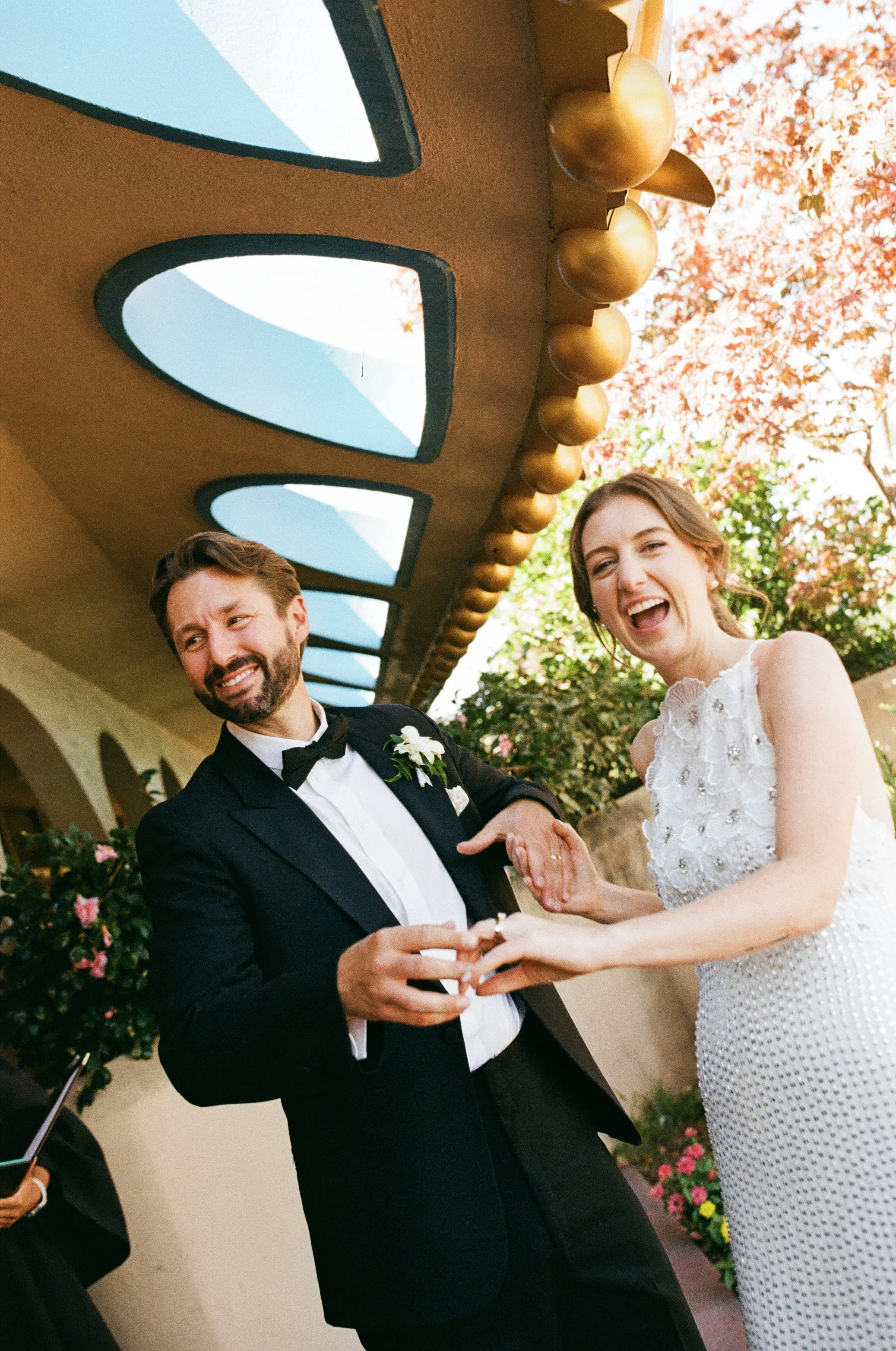 A couple dressed in formal wedding attire, smiling and holding hands during their wedding ceremony against a backdrop of pink flowers and autumn trees.