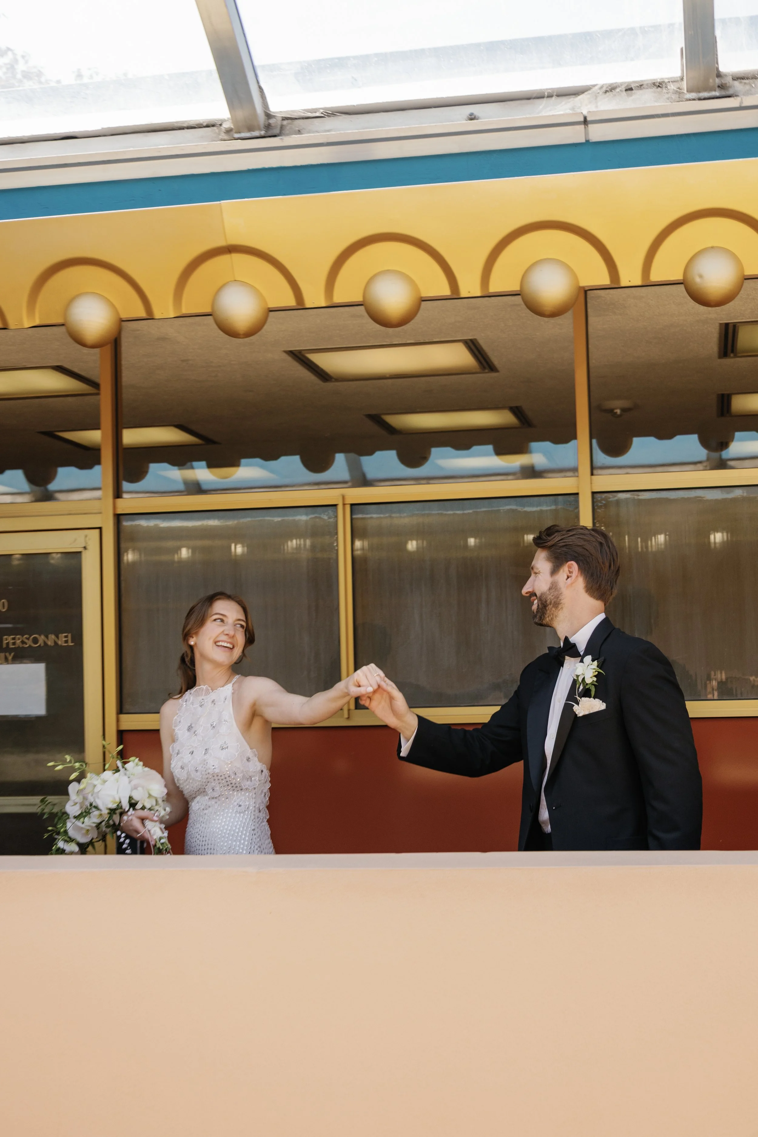 A bride and groom dancing outside a building with a yellow and blue facade, smiling and holding hands, wedding attire, wedding bouquet visible.