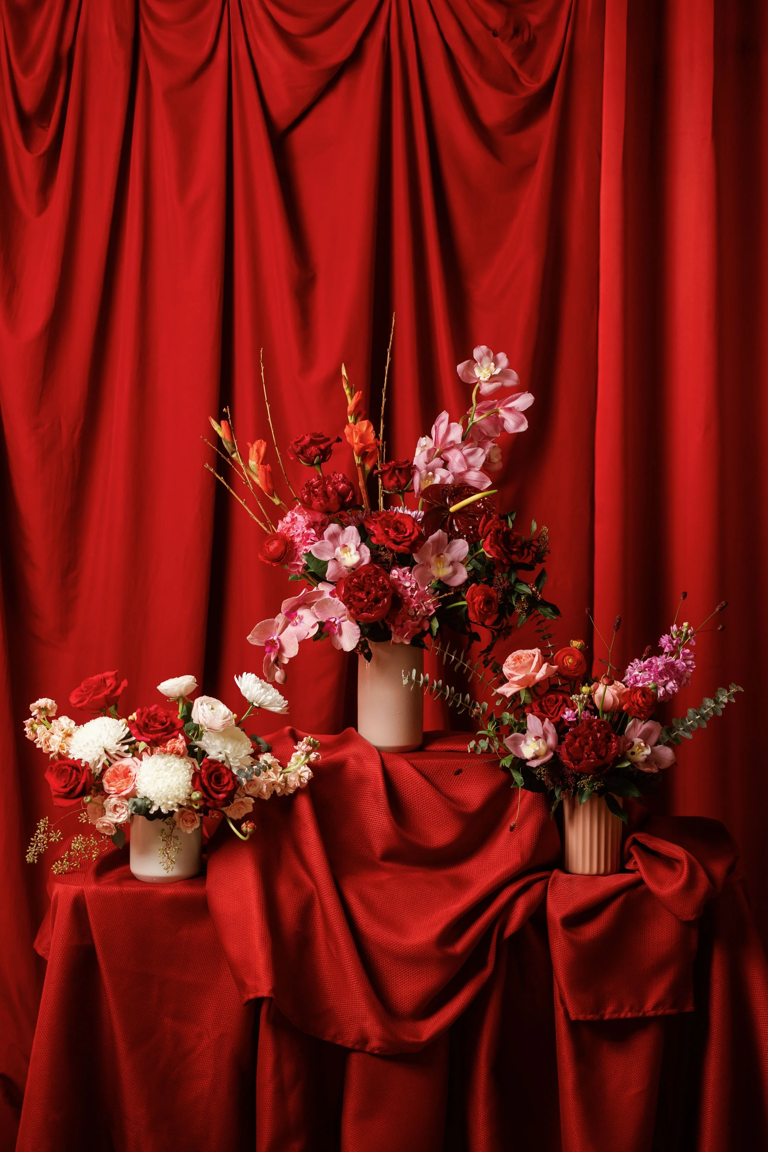 Arrangement of various flowers in white and peach vases against red velvet curtains.