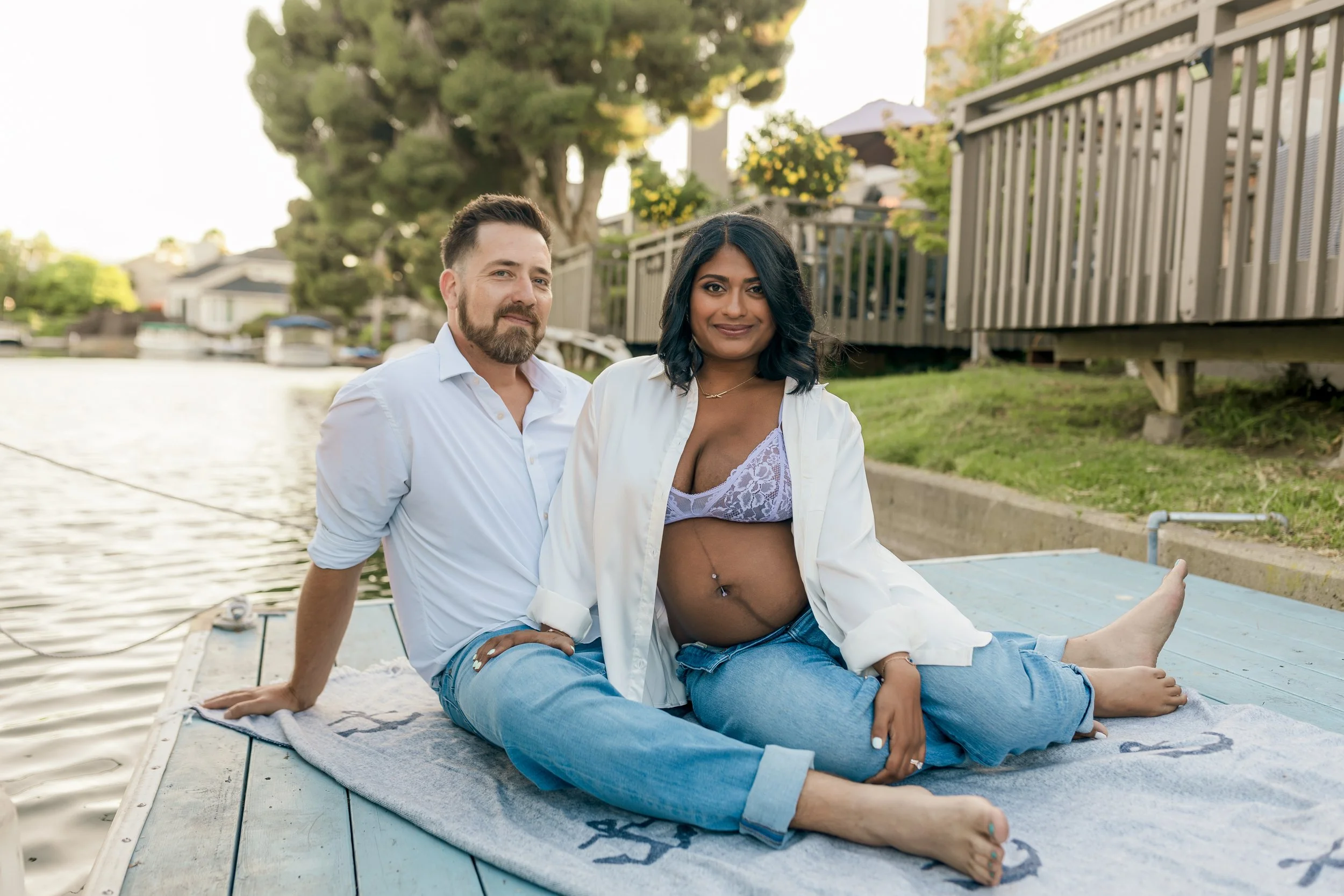 A couple by a waterfront, sitting on a dock with lush green trees and houses in the background during daytime.