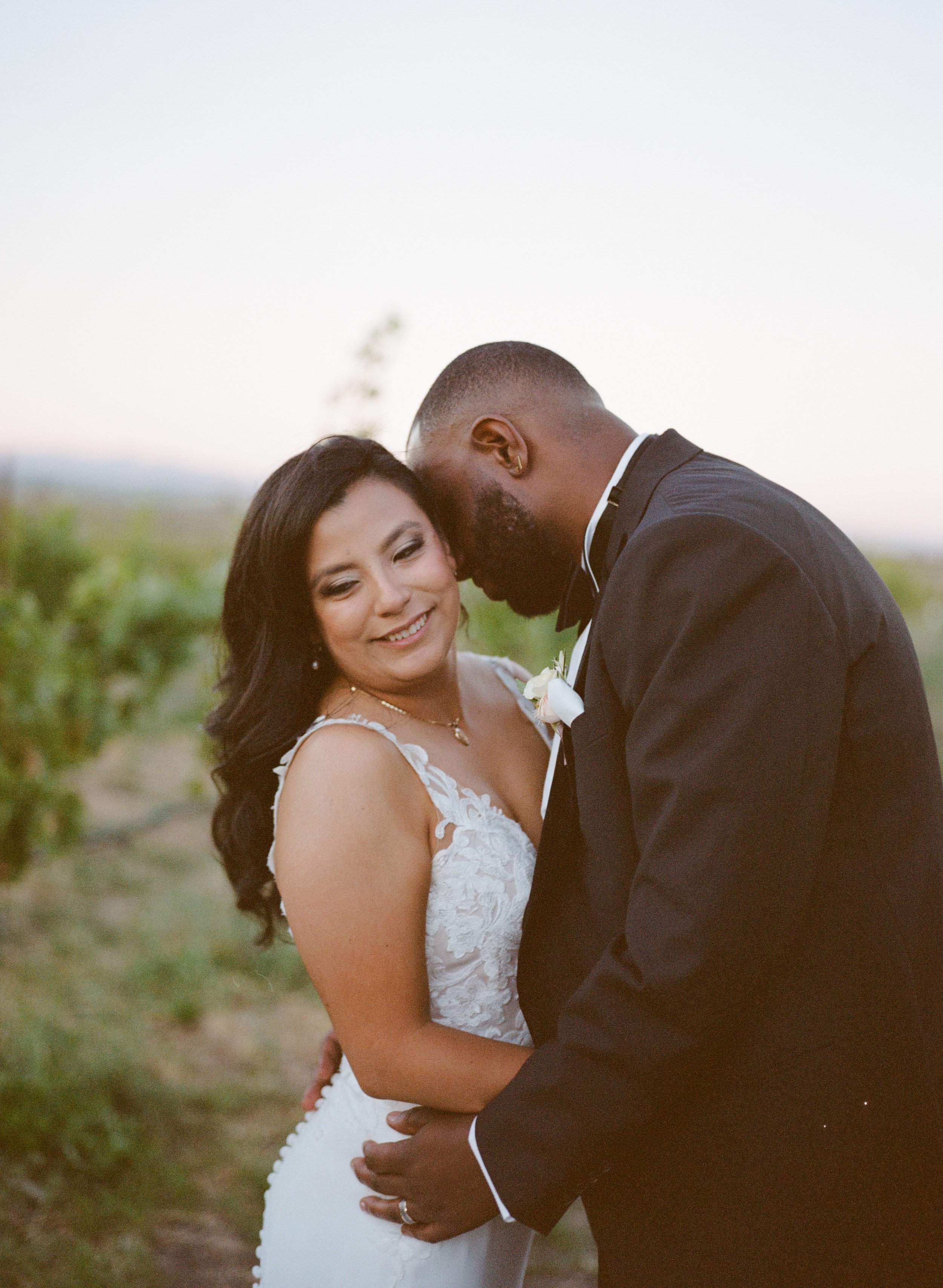 A bride and groom embrace outdoors during sunset, smiling and holding each other close.