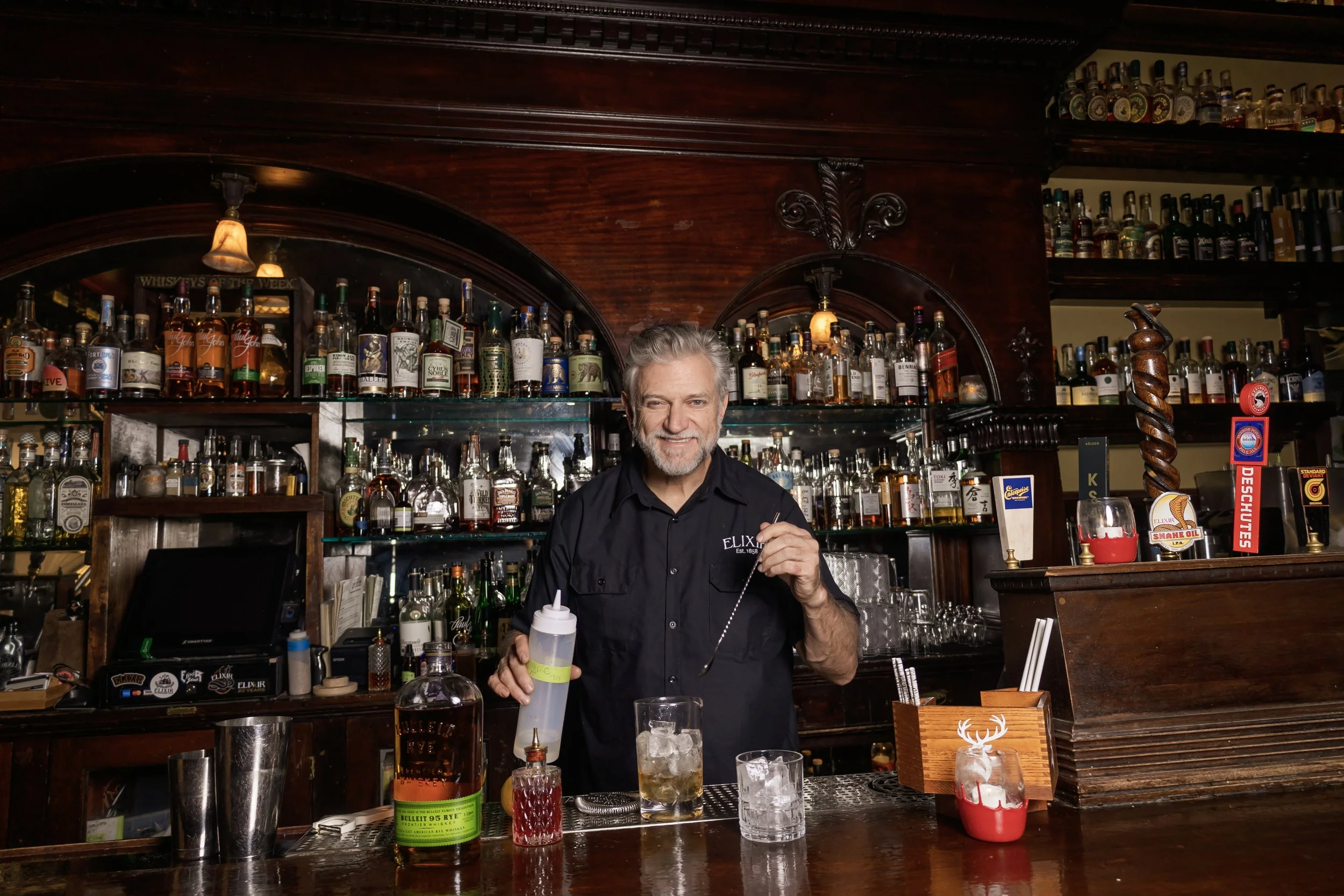 A bartender with gray hair and a beard smiles at the camera behind a bar, surrounded by numerous bottles of alcohol and bar paraphernalia.