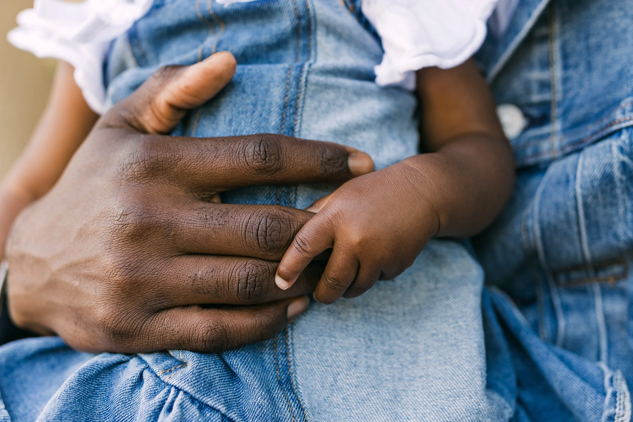 A close-up of a dark-skinned child's hand resting on an adult's hand against denim fabric clothing.