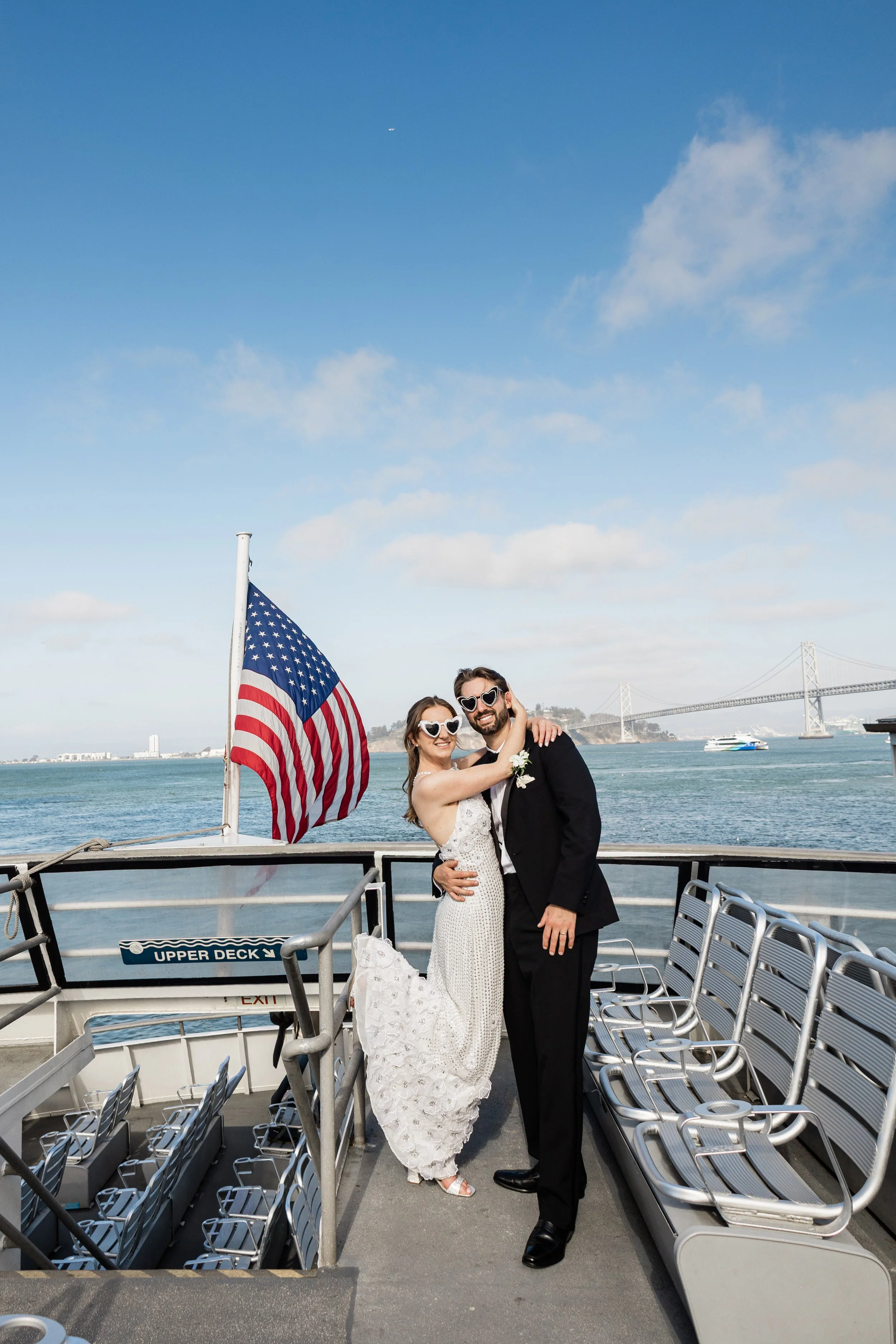 A newlywed couple on a boat deck, embracing and smiling. The bride is wearing a white lace wedding dress and heart-shaped sunglasses, and the groom is in a black tuxedo with heart-shaped sunglasses. An American flag is nearby, and water with a bridge