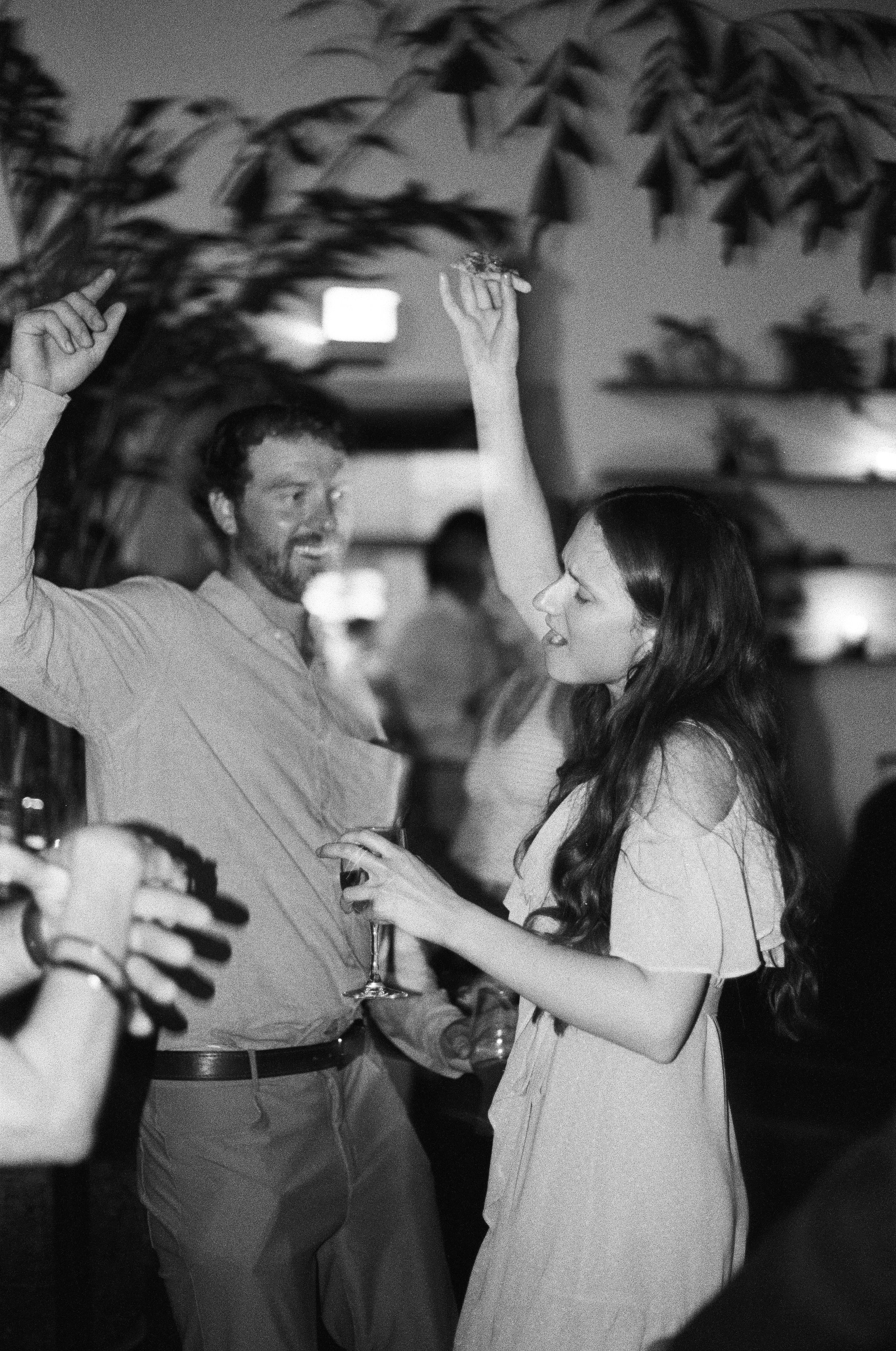 A man and woman enjoying a dance or conversation at a social event, with drinks in hand, in an indoor setting with string lights. The image is in black and white.