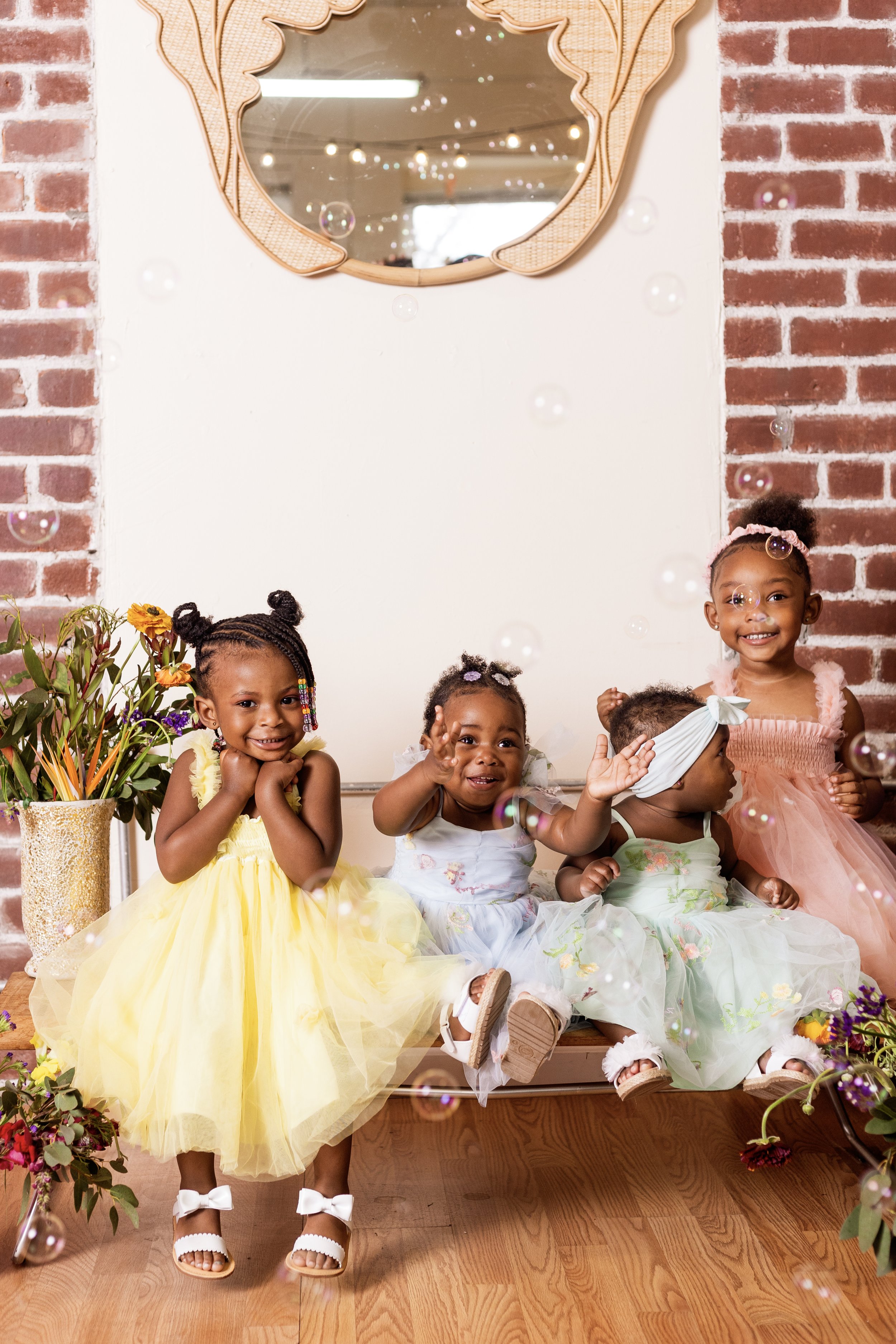 A group of young girls dressed in colorful dresses sitting on a bench with a brick wall and mirror behind them, surrounded by flowers and bubbles.