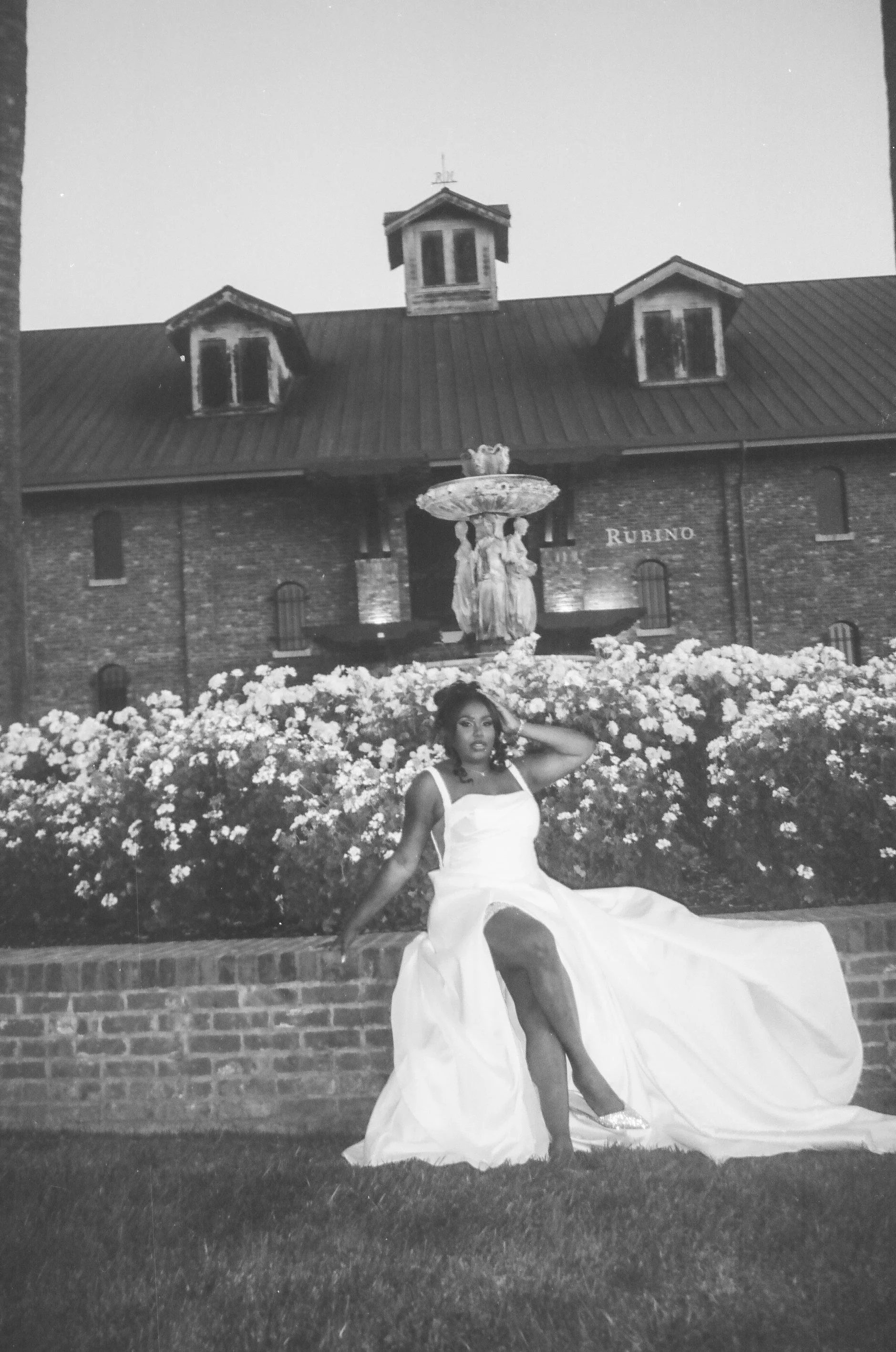 A woman in a wedding dress sitting on a brick ledge in front of a flower bed, with a building and a fountain in the background.