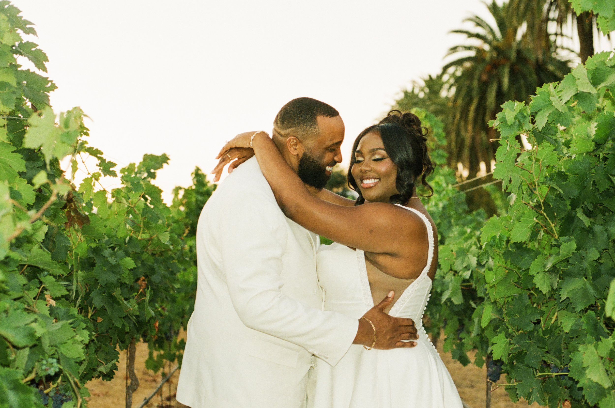 A couple dressed in wedding attire embracing outdoors surrounded by green vines and trees.