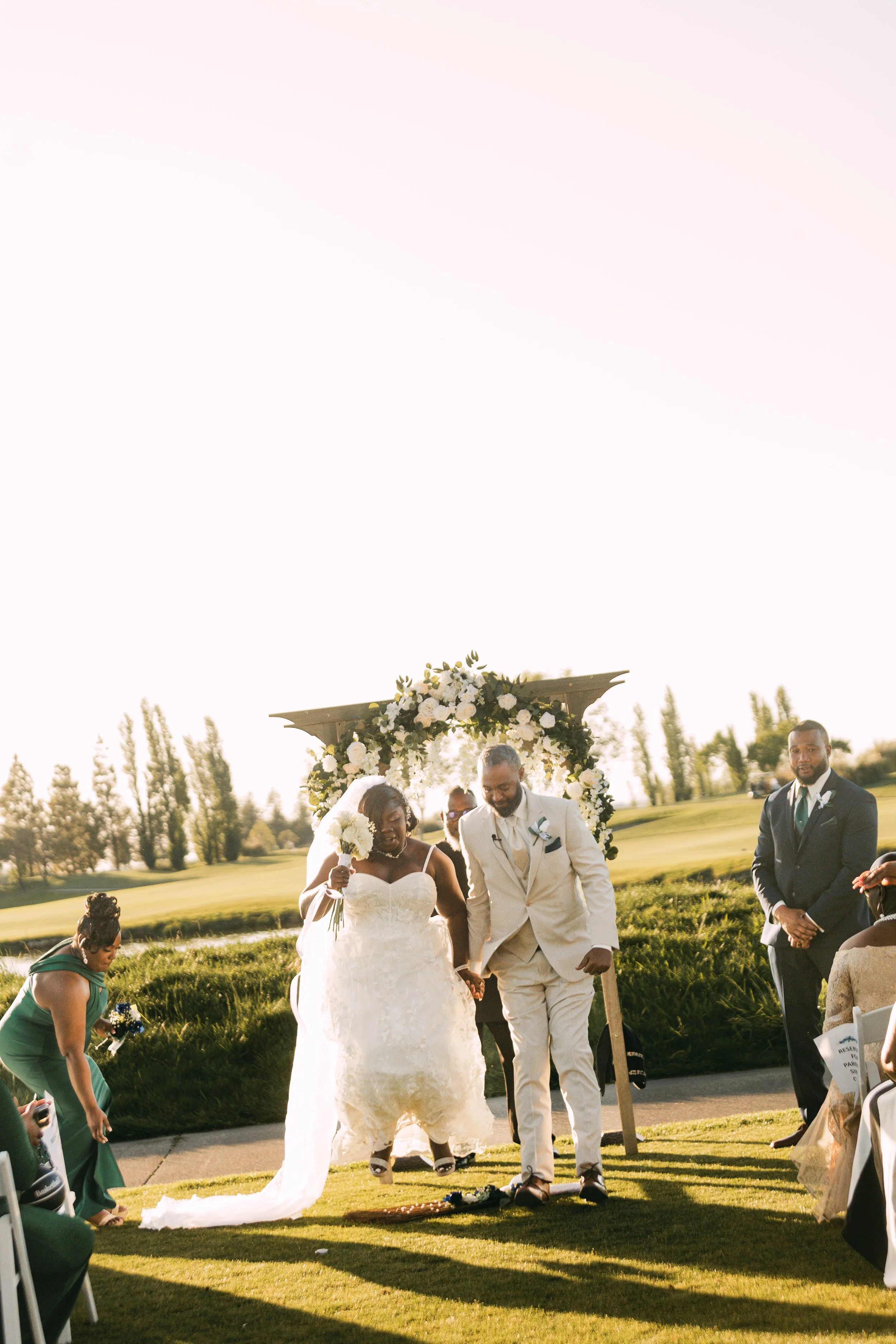 A wedding ceremony outdoors with a bride and groom walking arm in arm under a floral arch, surrounded by guests and greenery during sunset.