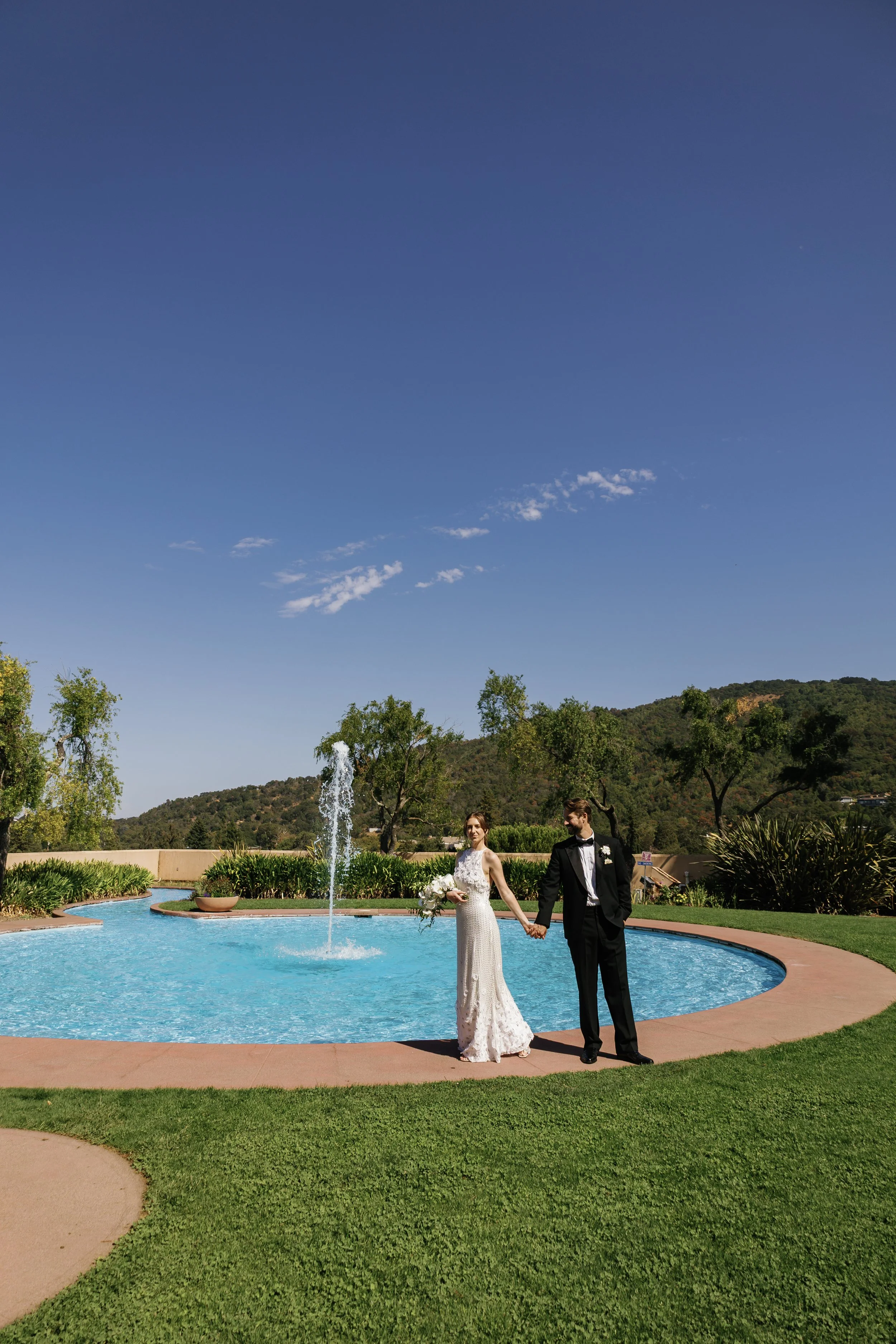A bride and groom walking hand in hand near a swimming pool, holding a bouquet, during their outdoor wedding ceremony on a sunny day with clear blue sky, trees, and hills in the background.
