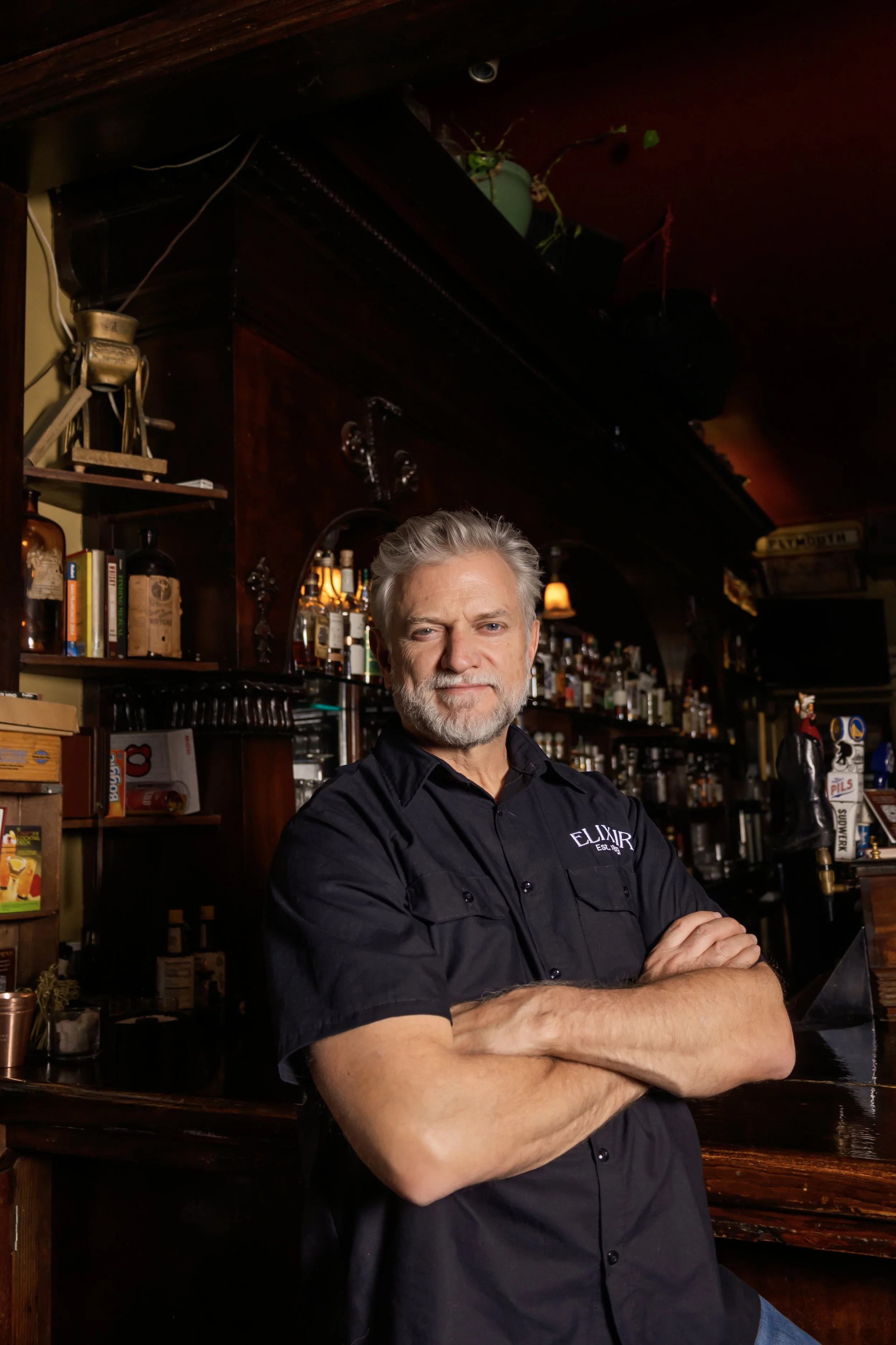 A man with gray hair and a beard standing with his arms crossed in a bar with shelves of liquor and books behind him.