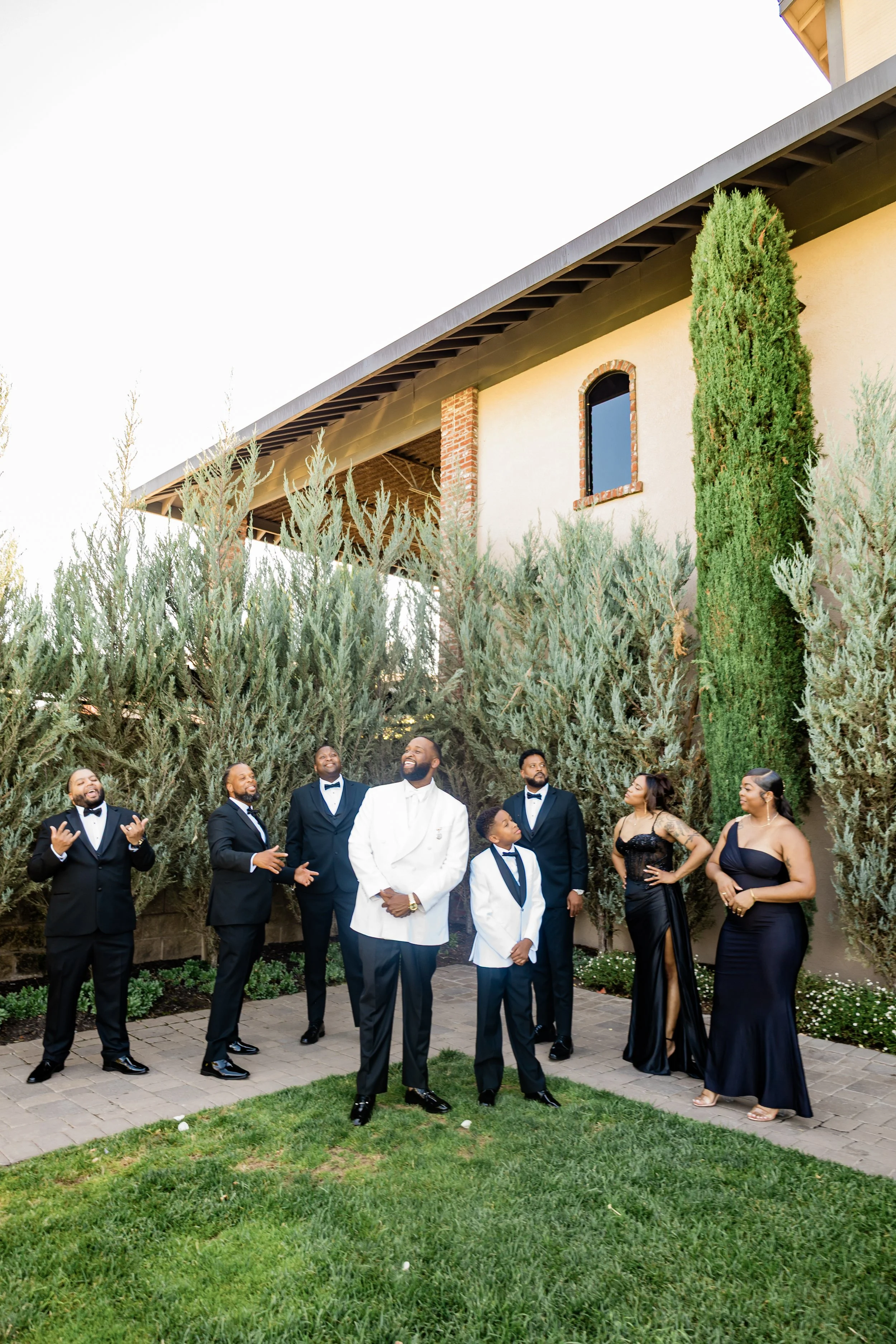 A group of nine people dressed in formal attire standing outdoors in front of tall bushes and a building. The men are wearing tuxedos or white jackets, and the women are in elegant black dresses. They appear to be enjoying a celebration or special ev