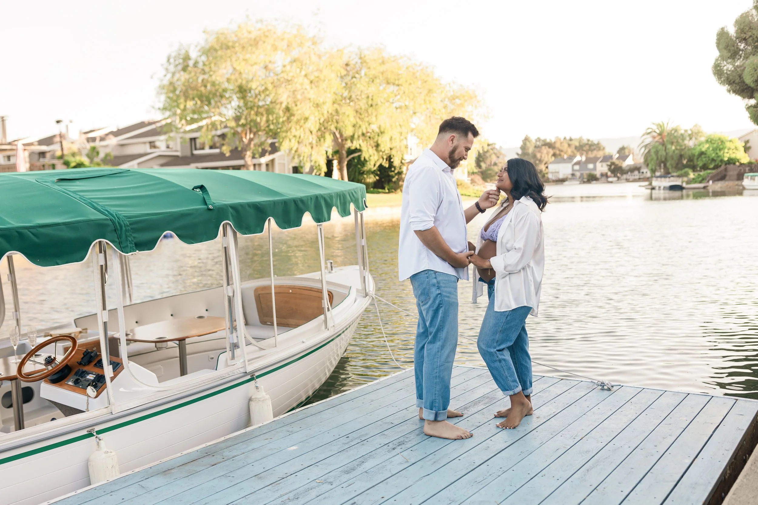 A man and woman rocking barefoot on a dock by a boat, with water and trees in the background, holding hands and smiling at each other.