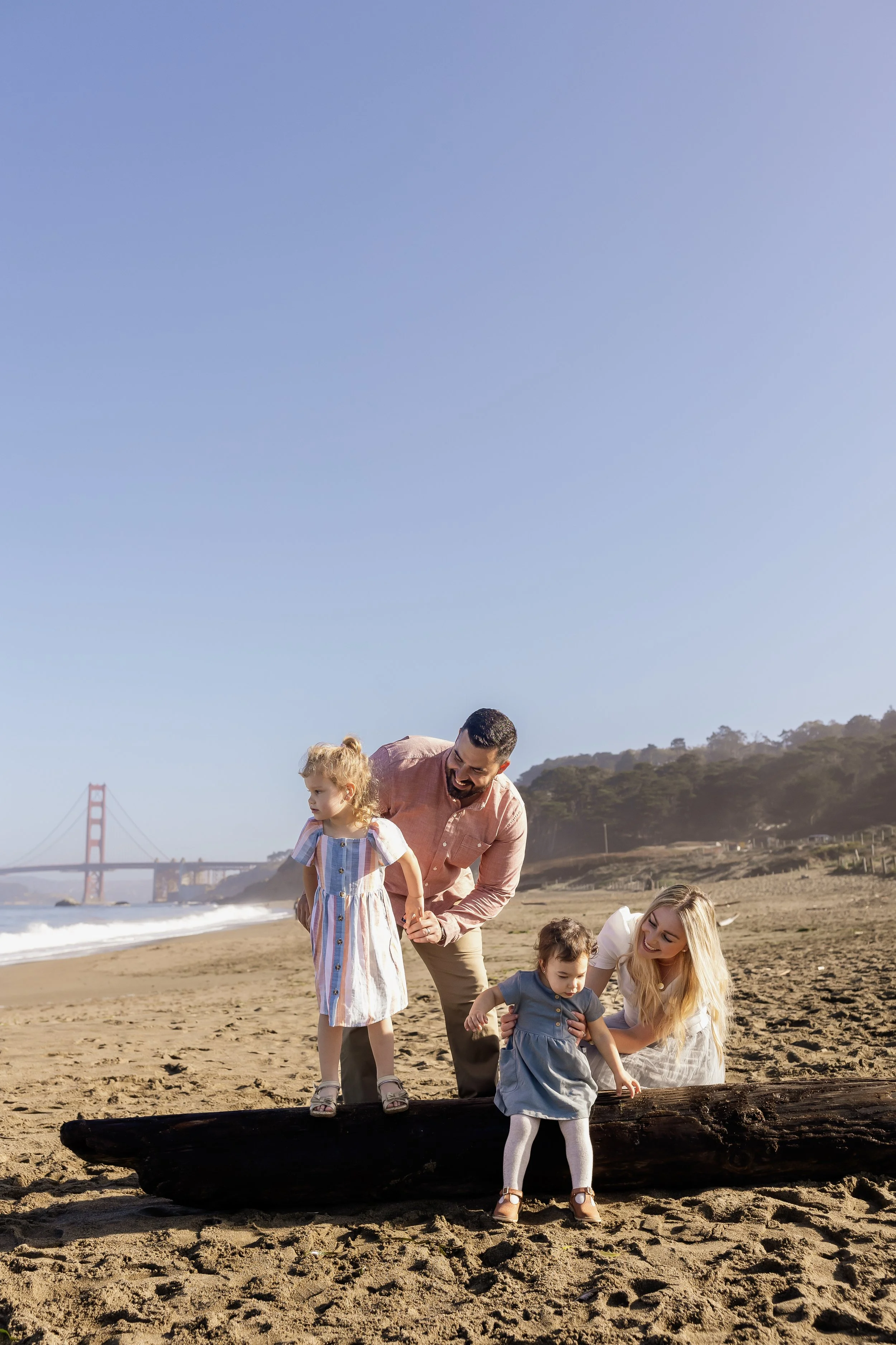 Family of five, including a man, a woman, and three young girls, enjoying time together on a sunny beach with the Golden Gate Bridge in the background. They are playing and exploring near a large driftwood log.