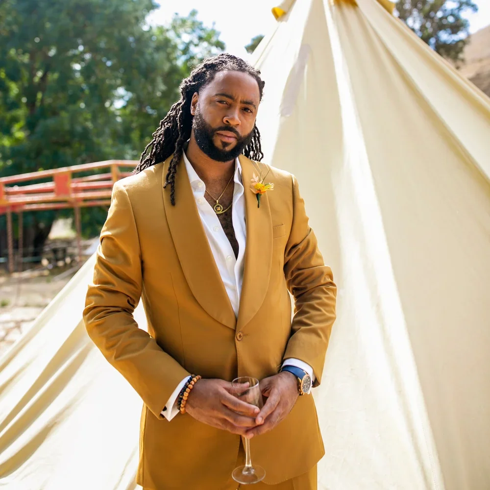 A man with dreadlocks wearing a gold suit, white shirt, and accessories stands outdoors holding a glass of wine, with a tent and trees in the background.