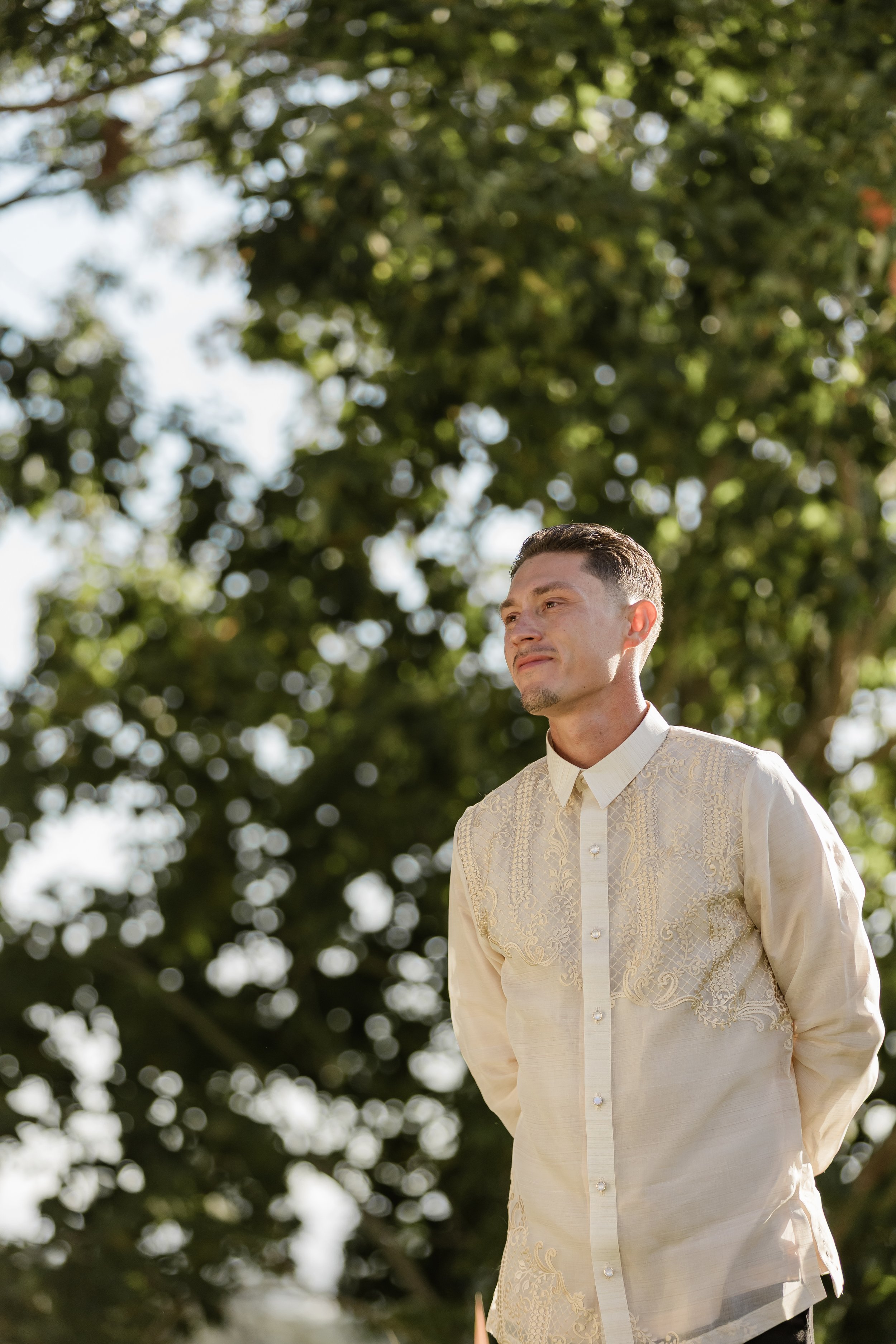 A young man wearing a traditional light-colored, embroidered barong tagalog stands outdoors in front of a lush green tree with sunlight filtering through the leaves.