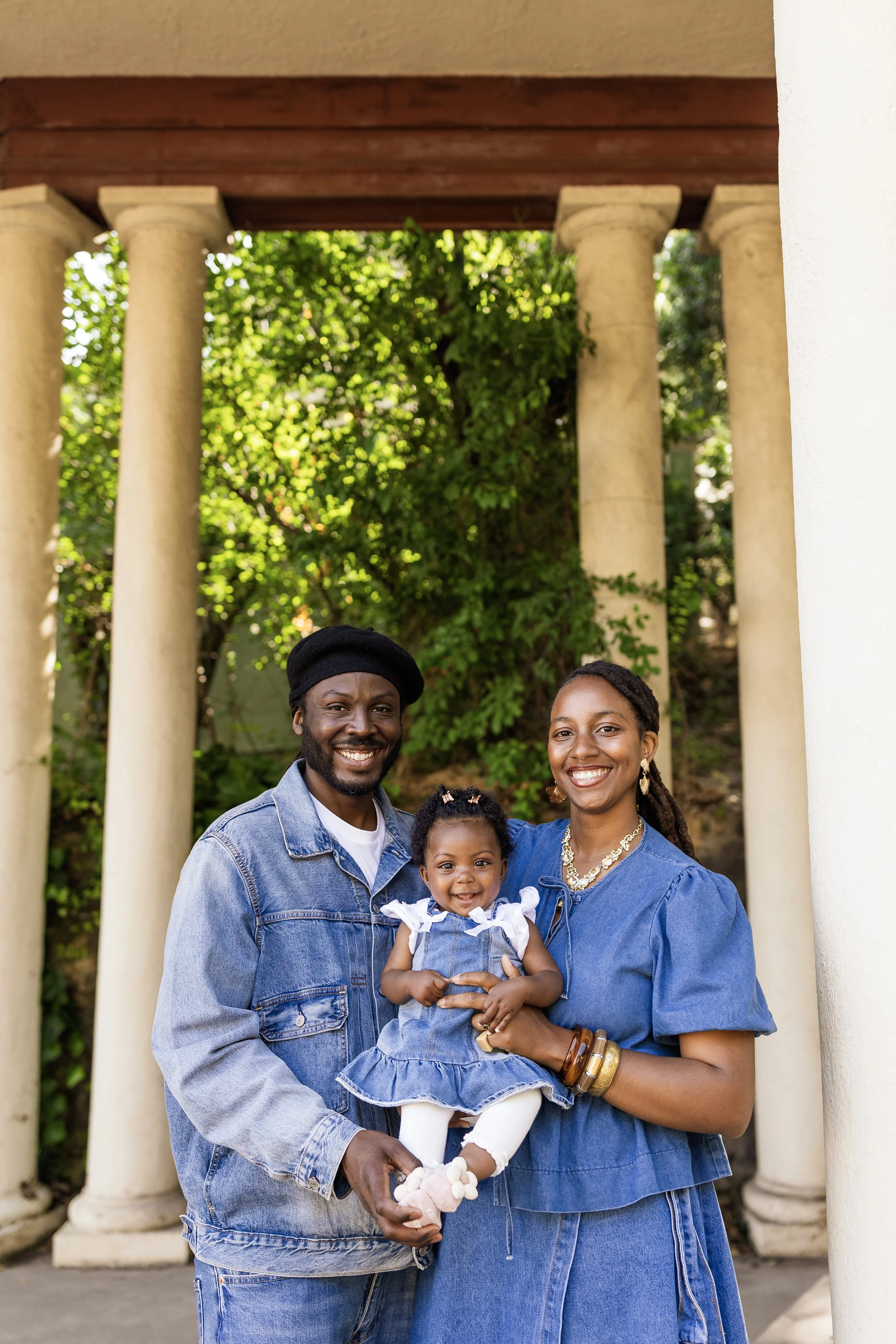 Family of three posing outdoors near white columns, green foliage background, man with beard, woman with jewelry, holding a smiling young girl in a denim dress.