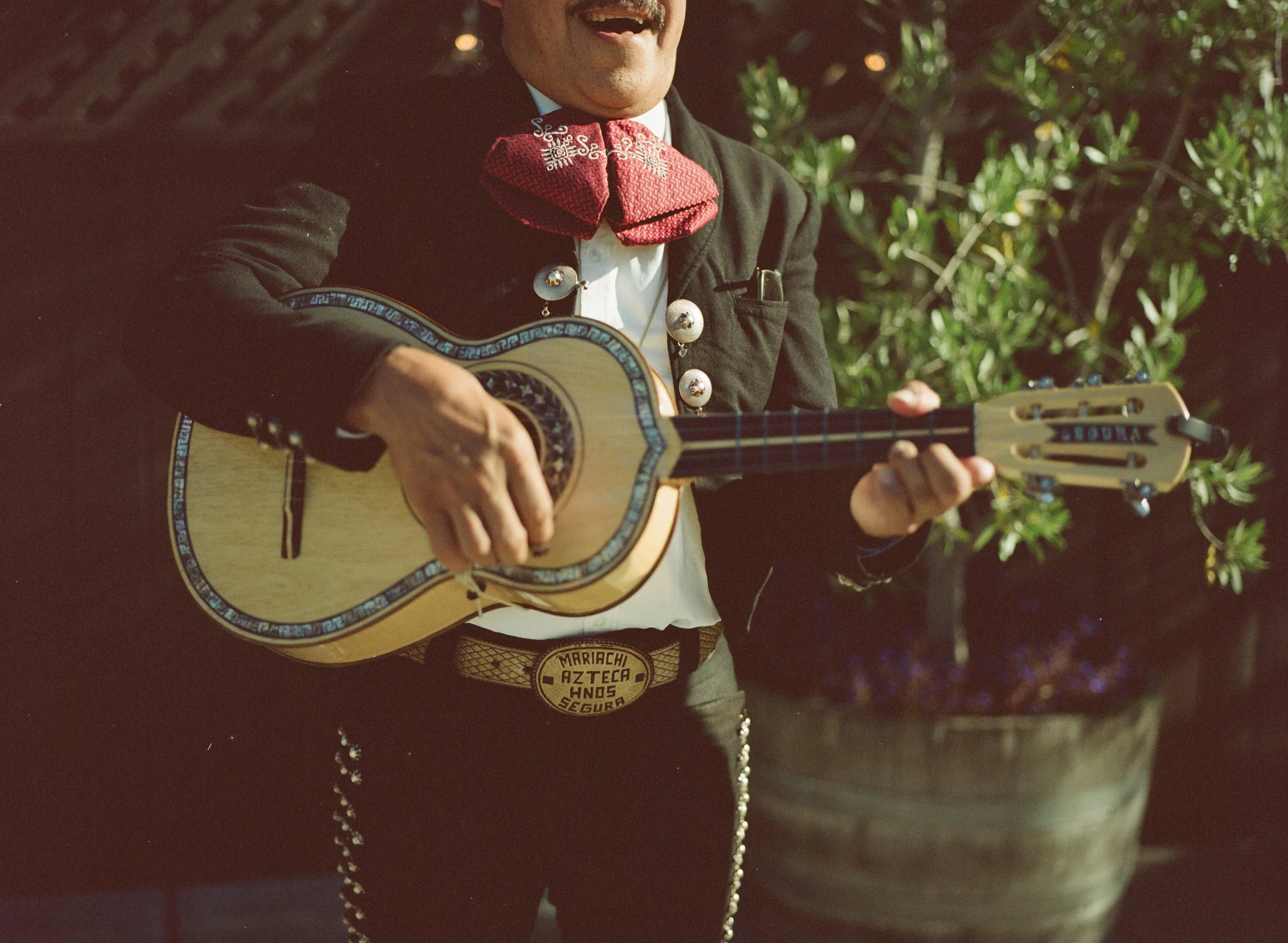 A person wearing a black jacket with silver buttons, a red bandana, and a bolo tie, playing an acoustic guitar outdoors in sunlight, with green foliage in the background.