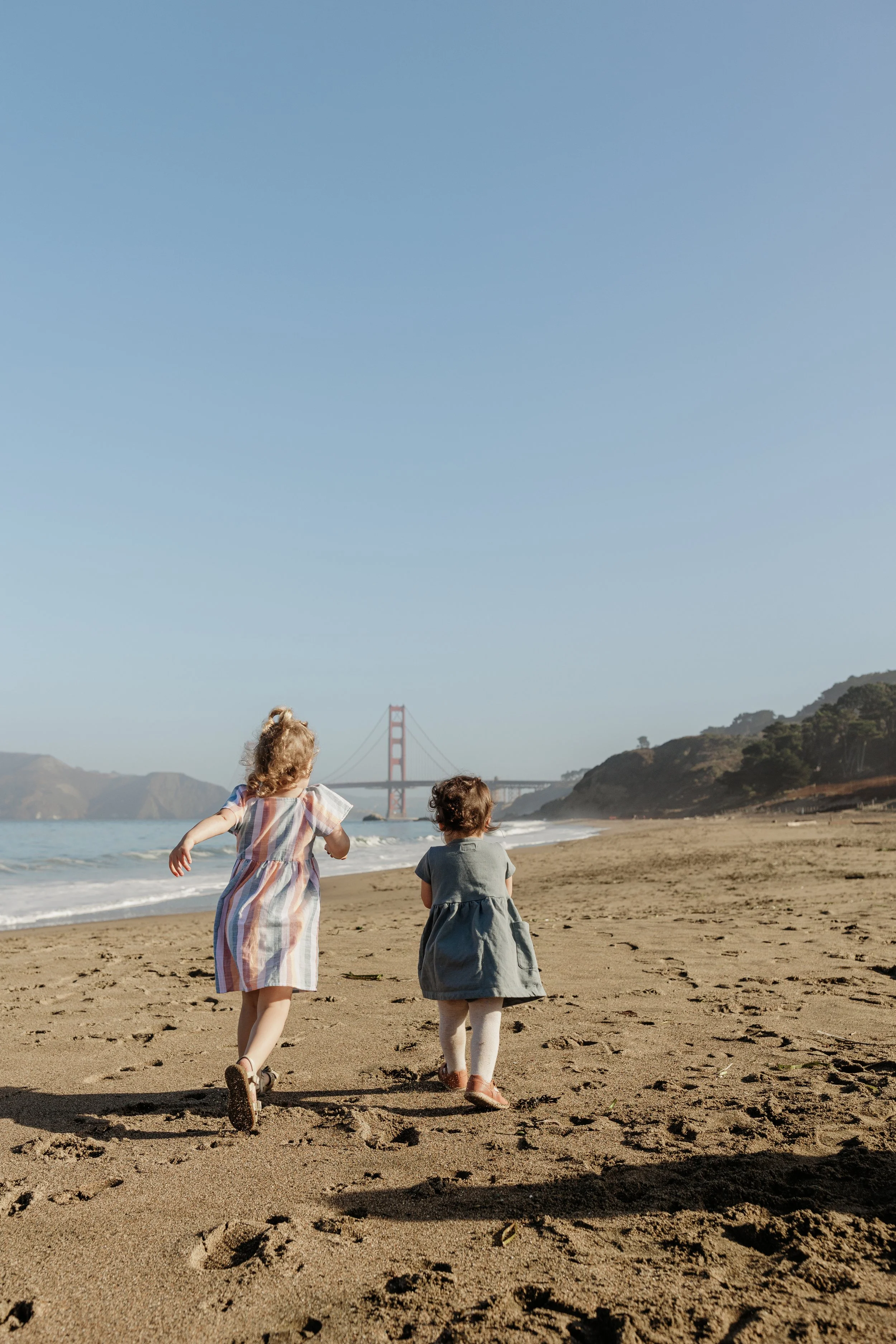 Two children running on a sandy beach with the Golden Gate Bridge in the background, hills, and a clear blue sky.