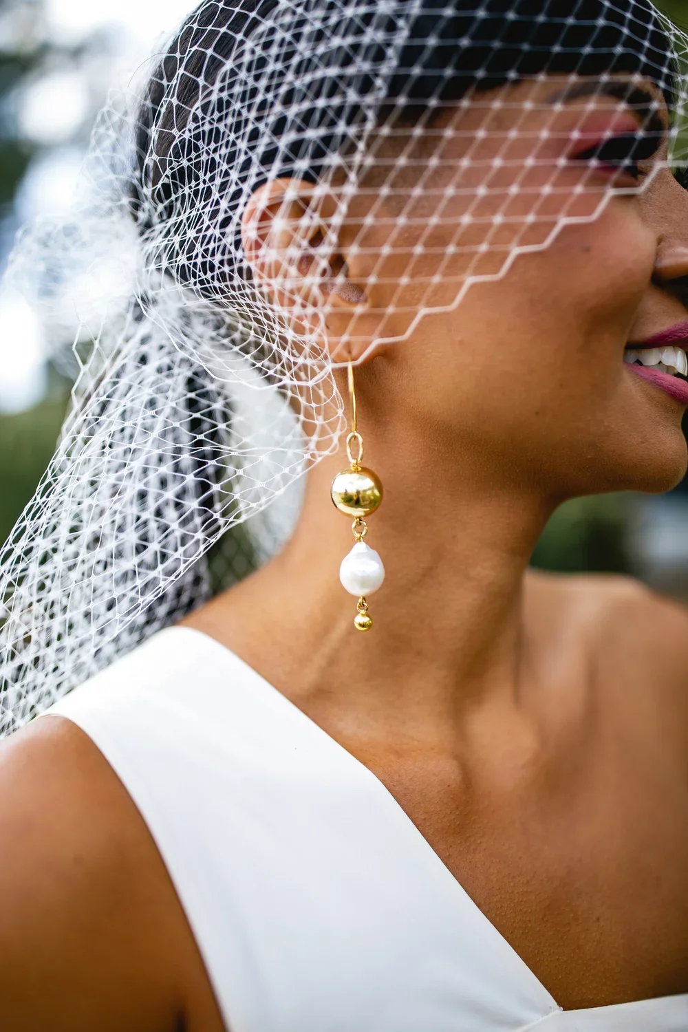 Close-up of a woman wearing a white dress and large gold and pearl earring, with a white netted veil over her face, smiling outdoors.