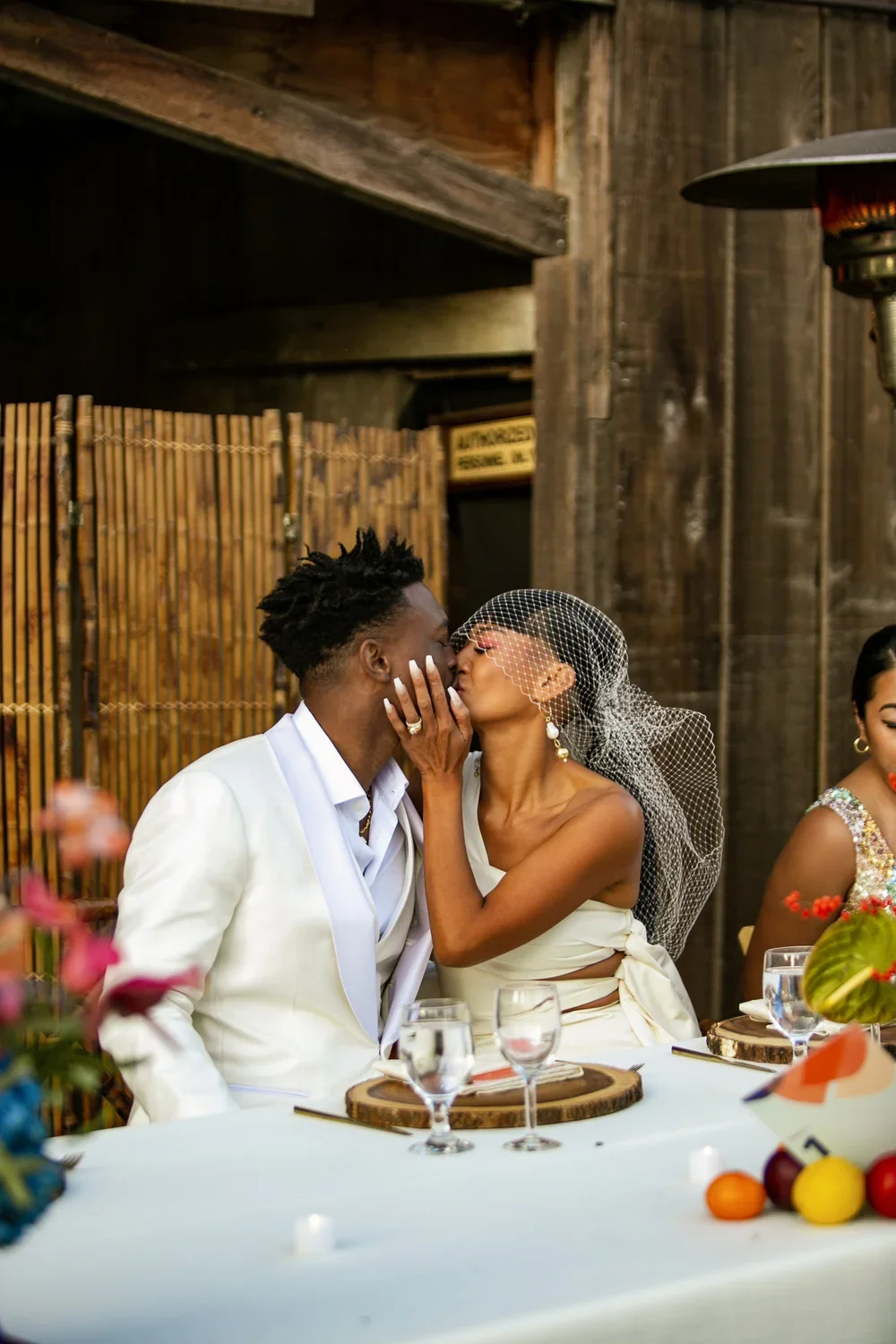 A bride and groom share a kiss at their wedding reception. The bride wears a white dress and veil, the groom wears a white suit. They sit at a decorated table with glasses and floral arrangements.
