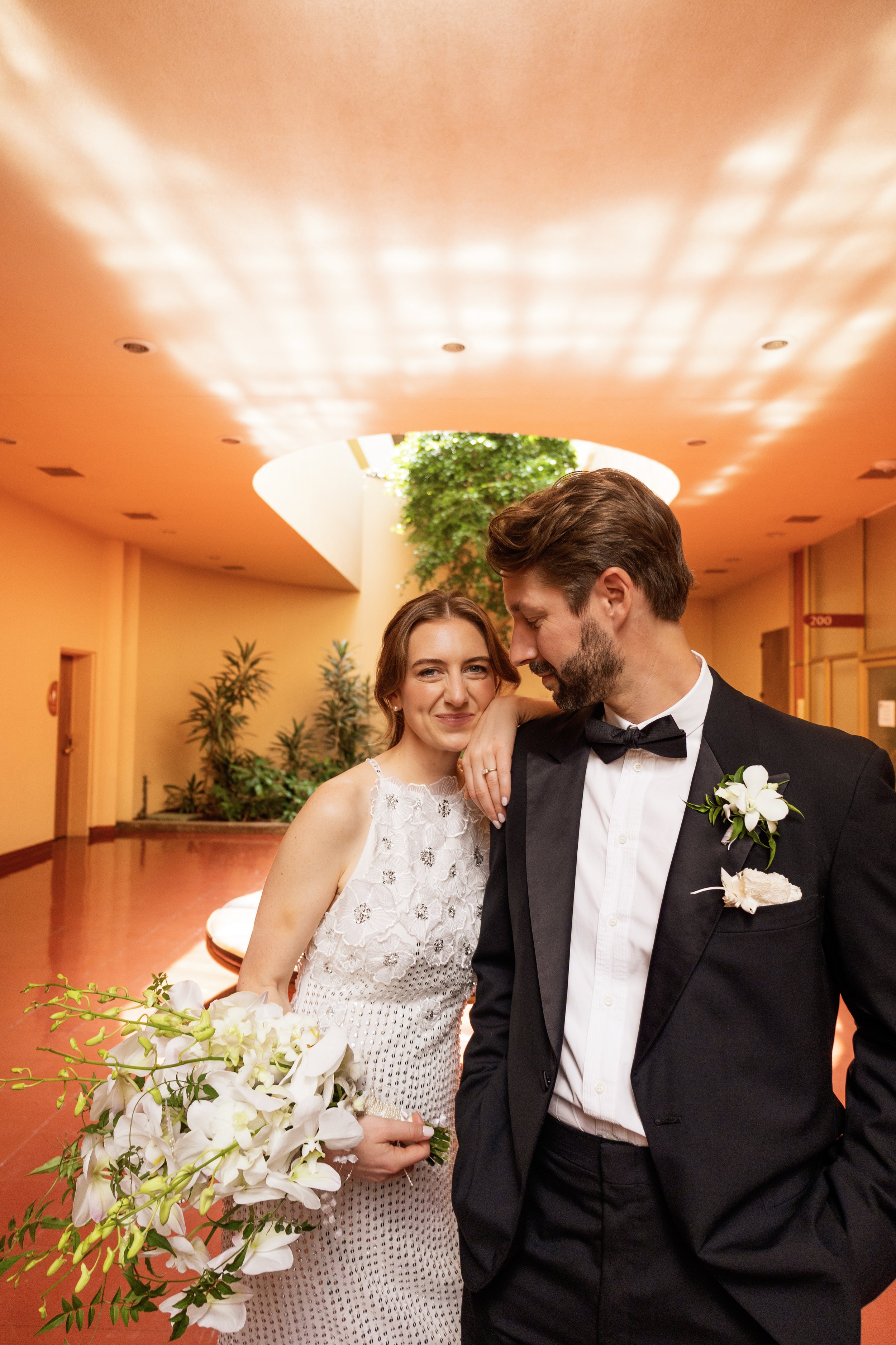 A bride and groom standing close together, smiling at each other in a warmly lit reception area with plants and a round table with white flowers in the foreground.