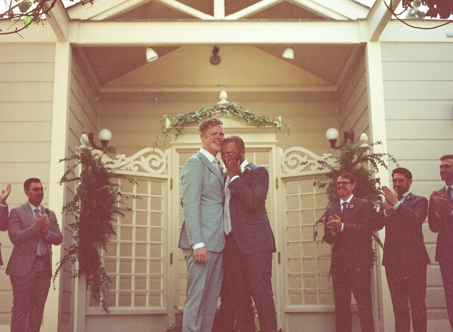 A couple in wedding attire sharing a moment with a group of groomsmen or friends clapping around them at an outdoor wedding ceremony.