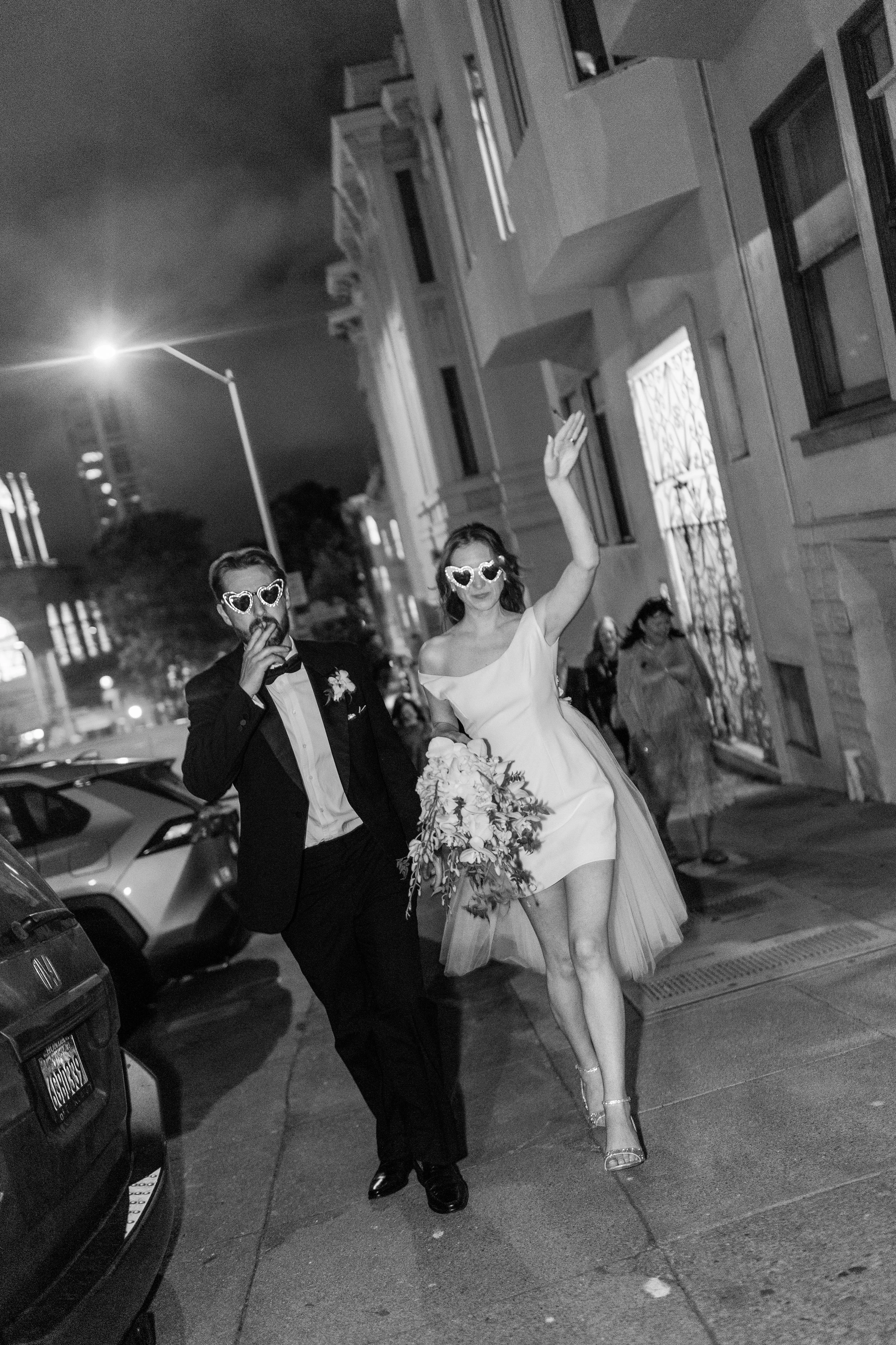 A bride in a white dress and heels walking with a groom in a tuxedo during a nighttime outdoor wedding, both wearing heart-shaped sunglasses, surrounded by guests and parked cars on a city street.